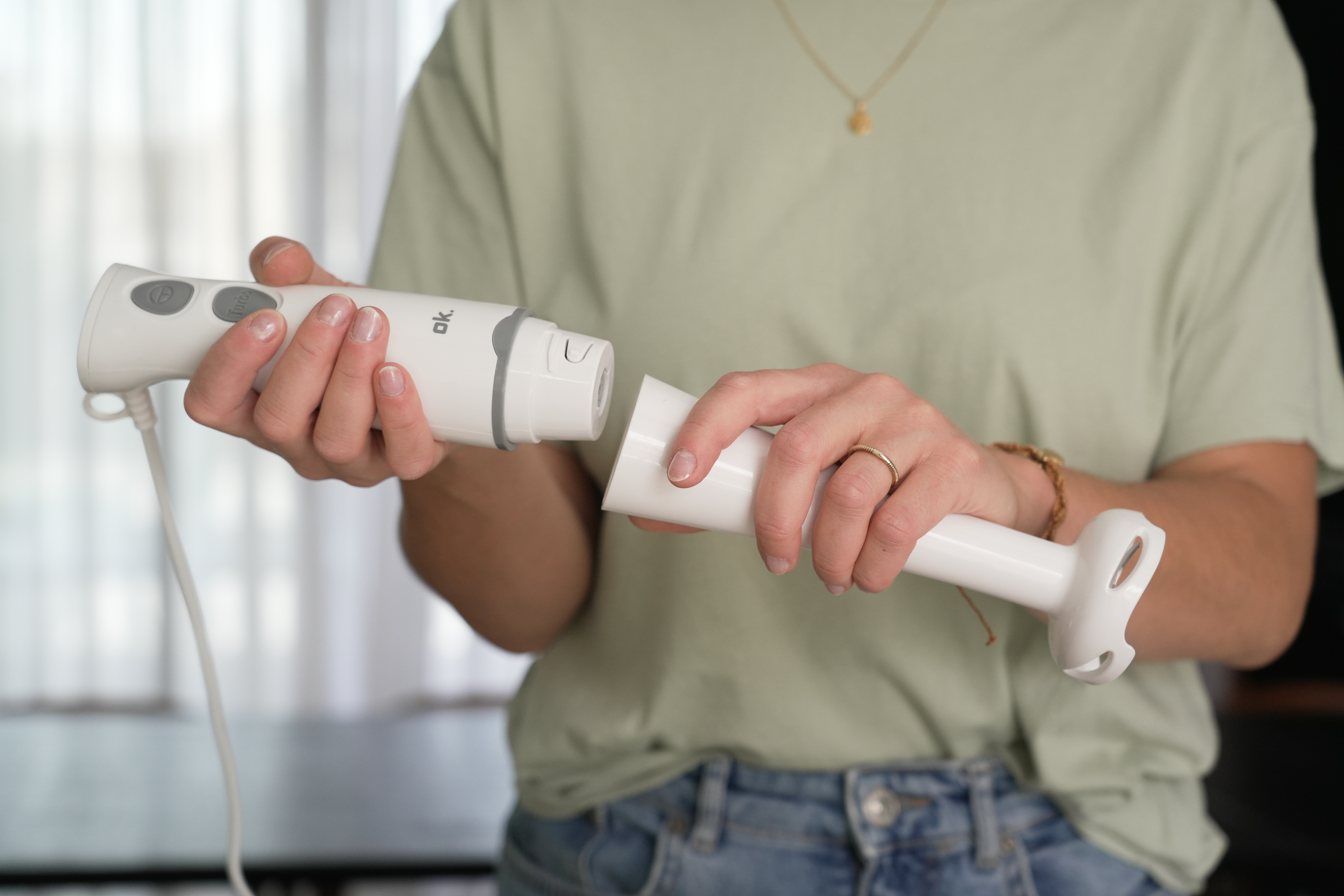 Osoba montuje biały blender. Ręce trzymają części na tle jasnozielonej koszuli i dżinsów.