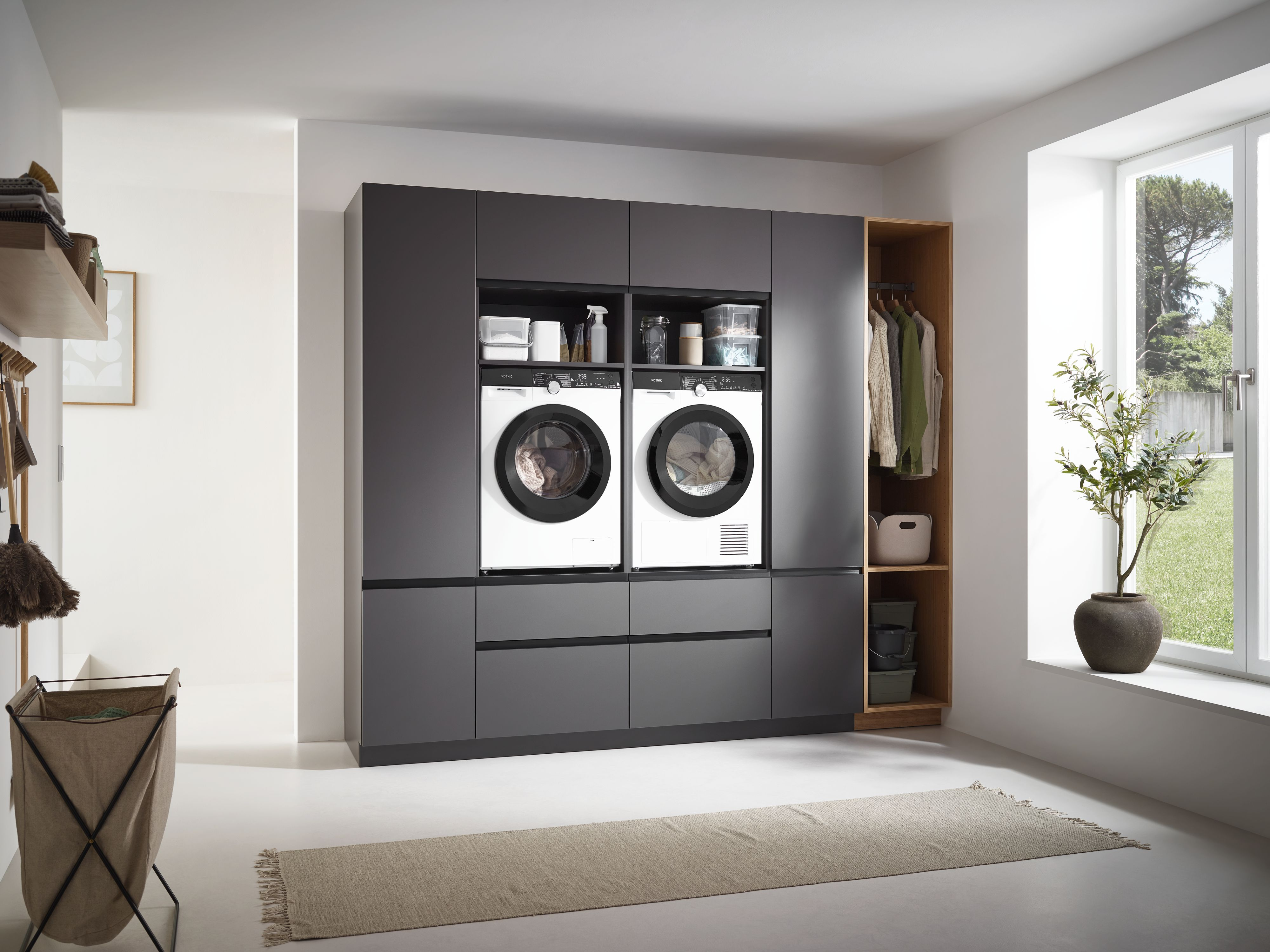 Waschküche mit Waschmaschinen, Trockner und Schränken. Kleidung hängt in einem Holzschrank. Rechts ist ein Fenster.