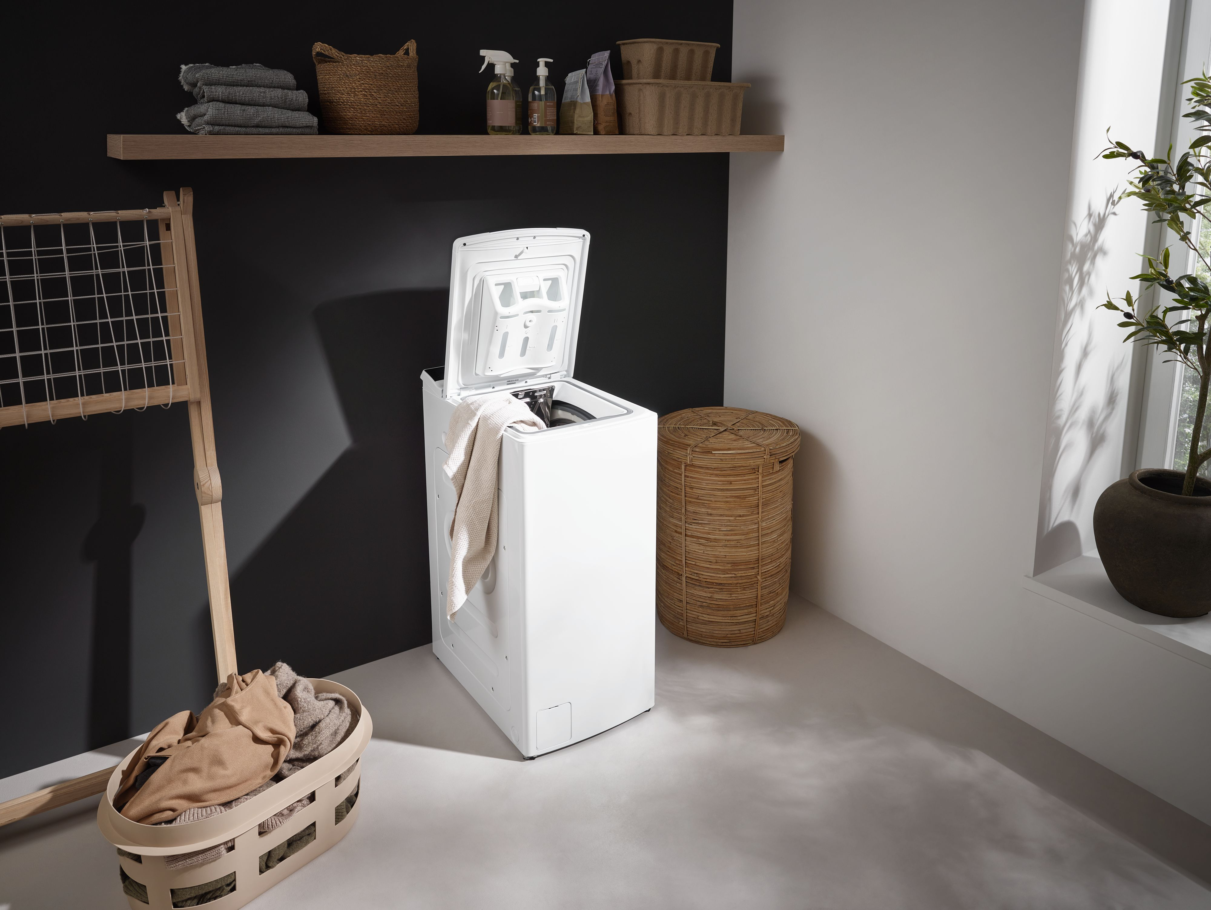 Eine weiße Waschmaschine mit offenem Deckel, Wäschekorb und einem Holzregal mit Gegenständen. Eine Pflanze steht in der Ecke.