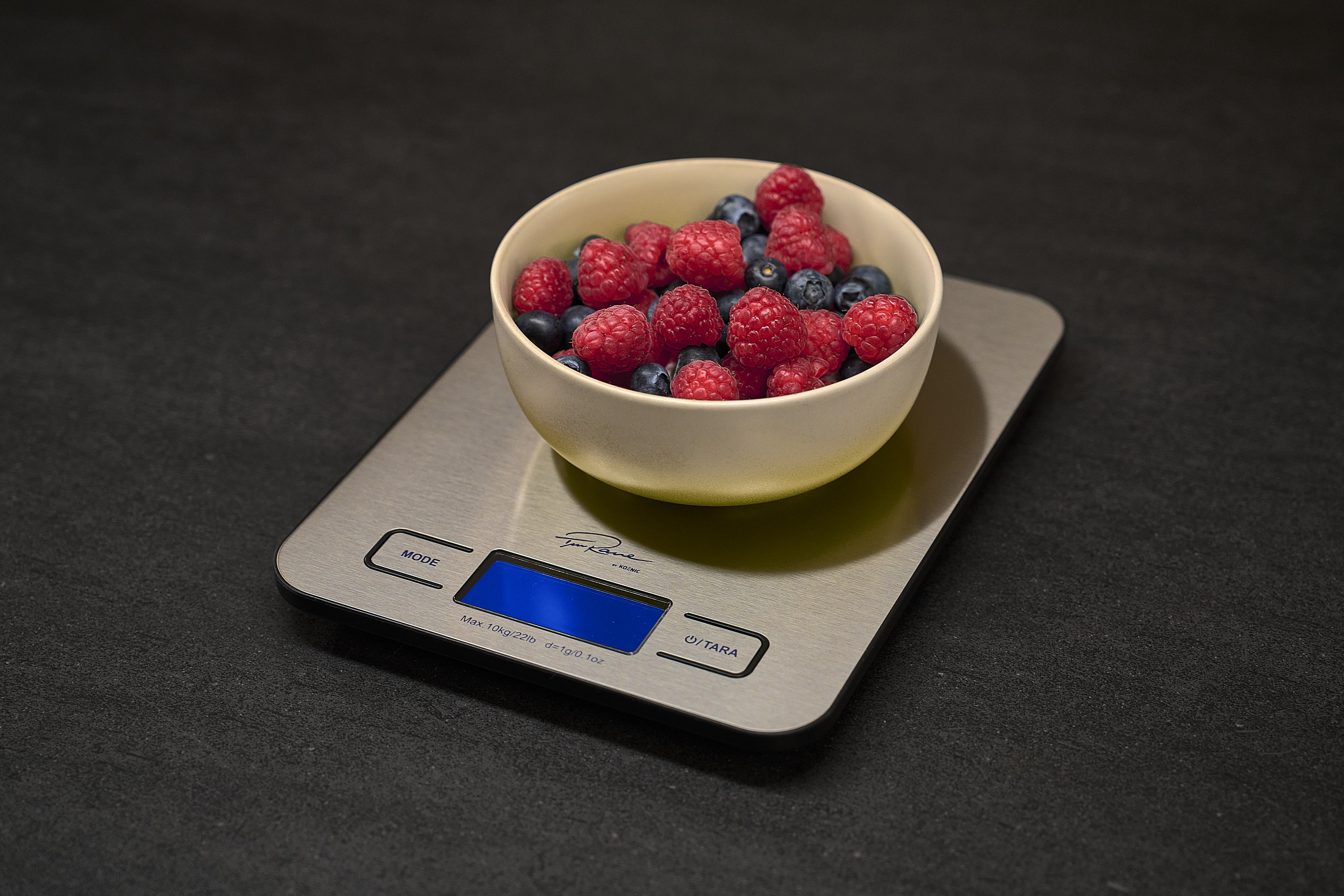 Eine Schüssel mit Himbeeren und Blaubeeren steht auf einer digitalen Küchenwaage. Die Waage steht auf einer dunklen Oberfläche.