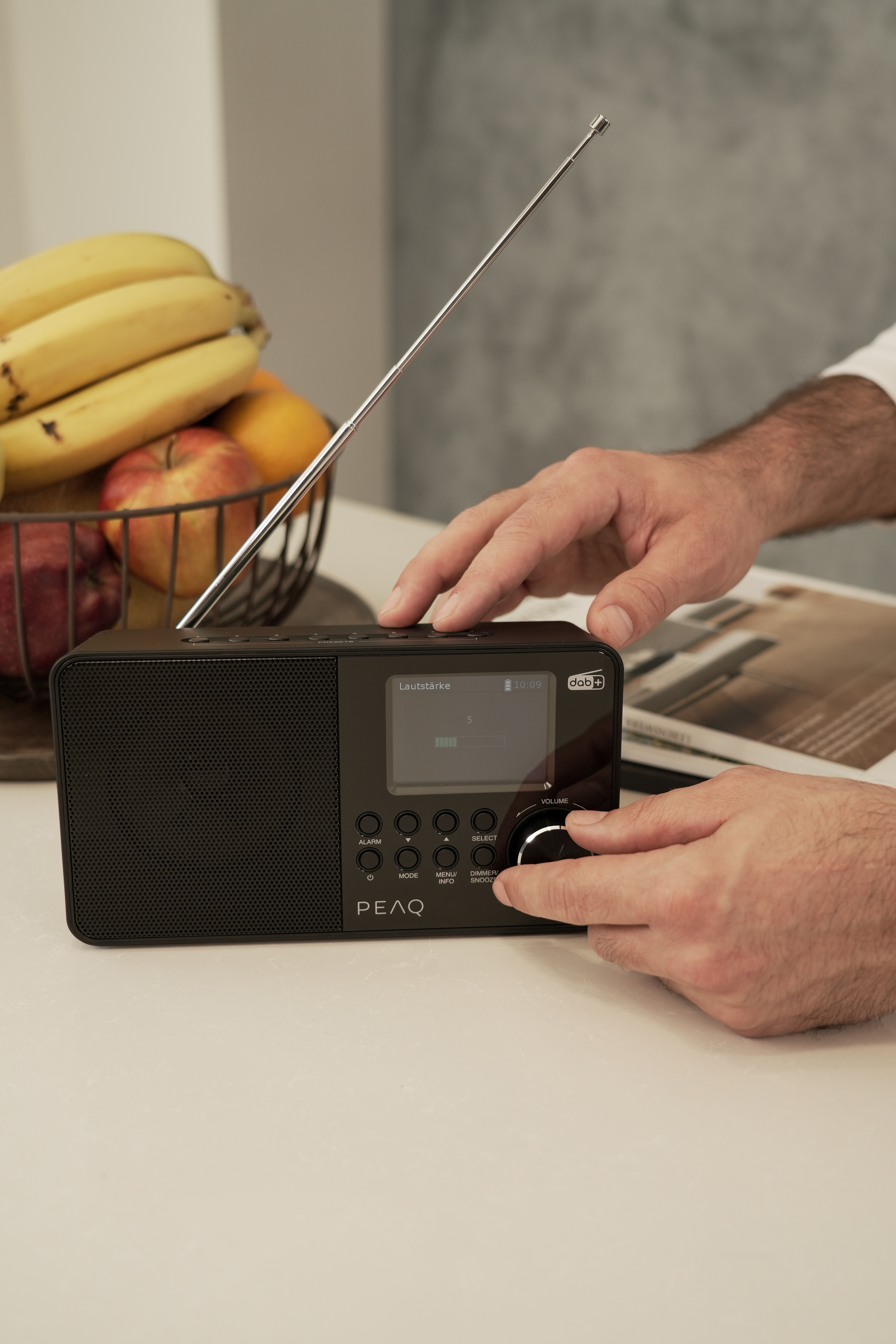 Ręka reguluje pokrętło głośności czarnego radia z cyfrowym wyświetlaczem. Kosz owoców w tle.