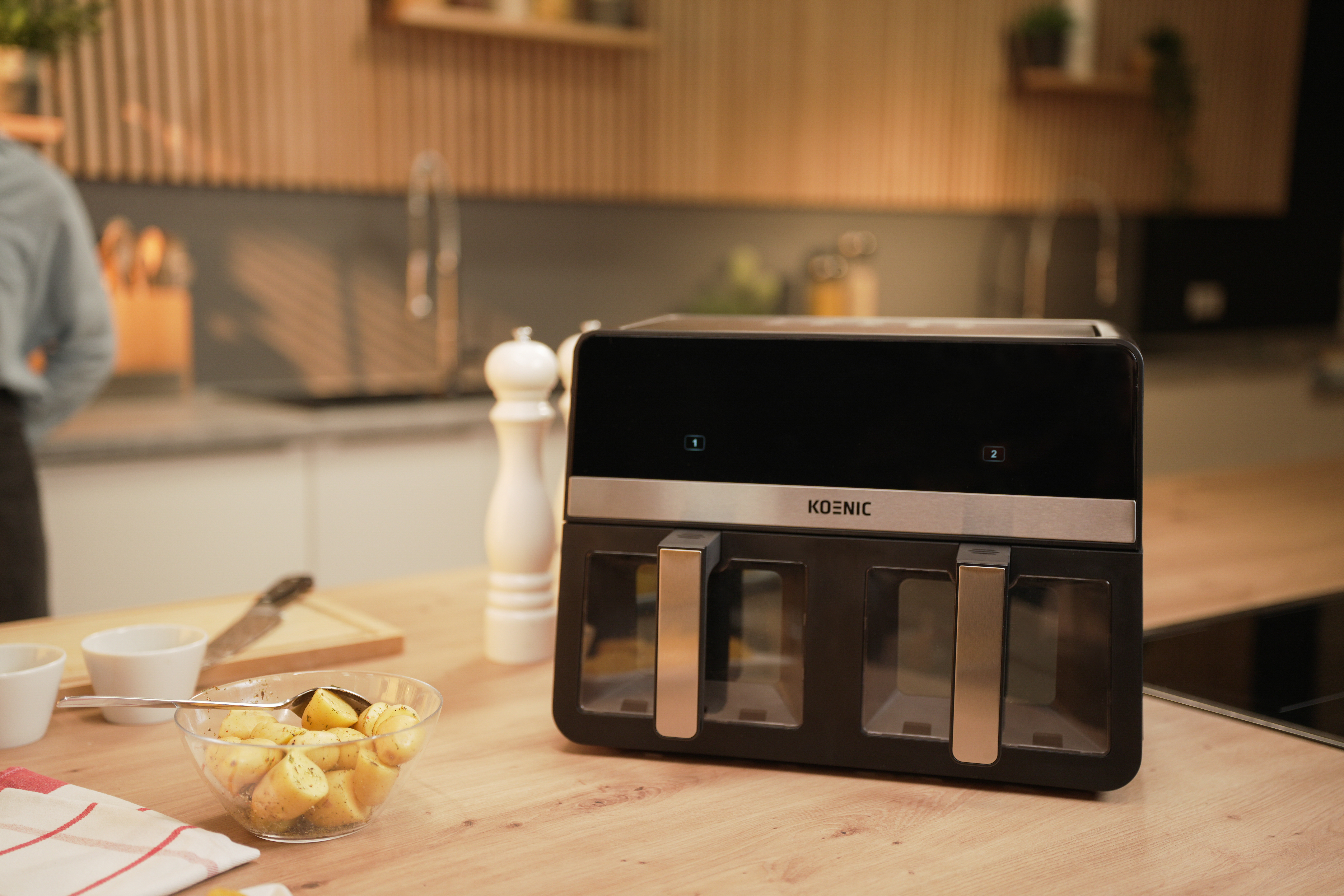 Frytownica powietrzna na drewnianym blacie z miską ziemniaków. Za nią stoi osoba. Kuchnia w tle.