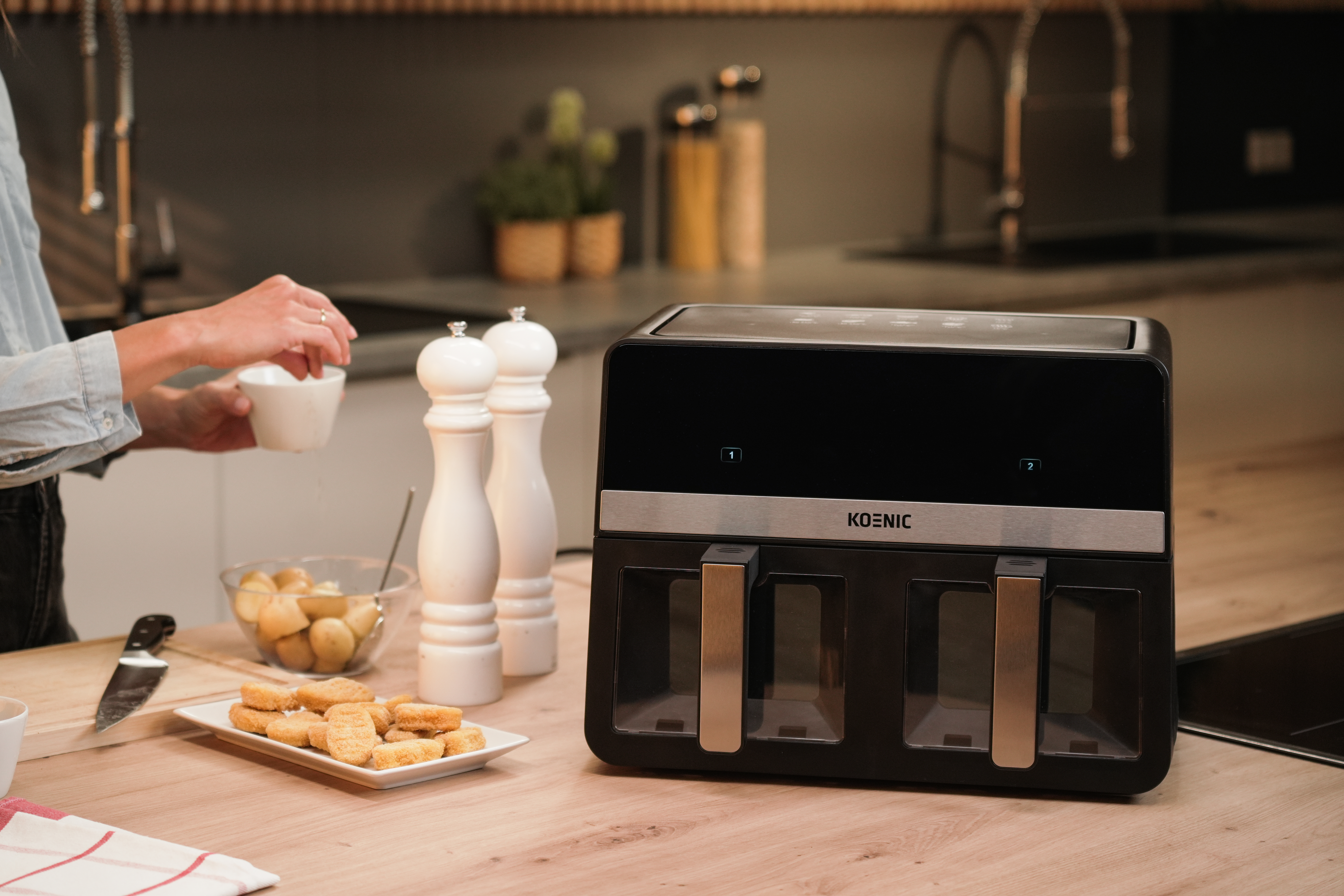 Scena kuchenna z czarną frytkownicą powietrzną, składnikami i osobą trzymającą filiżankę. Jedzenie jest na talerzu.