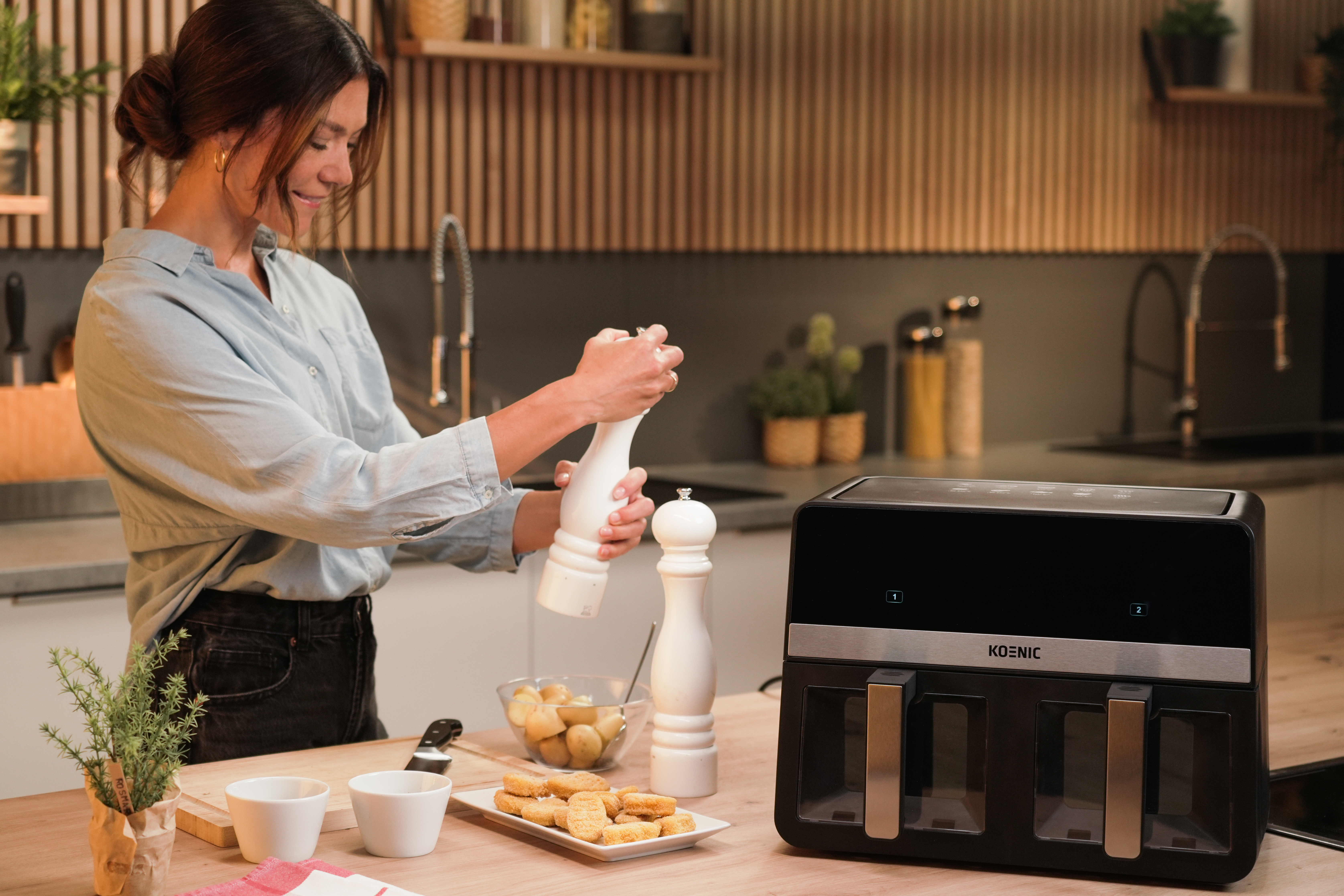 Kobieta w kuchni mieląca pieprz. Frytkownica beztłuszczowa, jedzenie i rośliny na blacie. Drewniane szafki w tle.