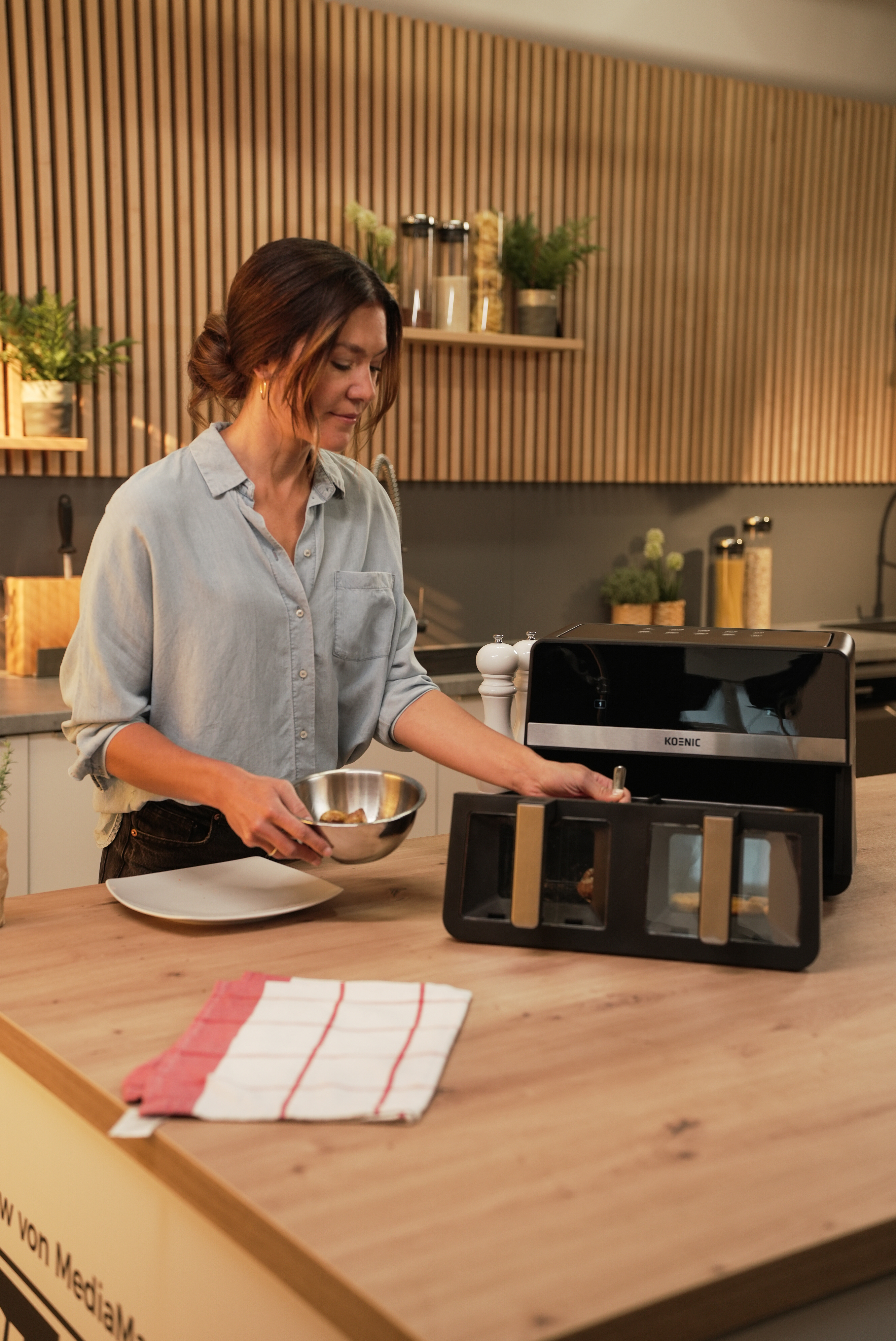 Kobieta wkłada jedzenie do tostera. Tło kuchni z drewnianymi panelami. Talerz, miska i ręcznik na blacie.