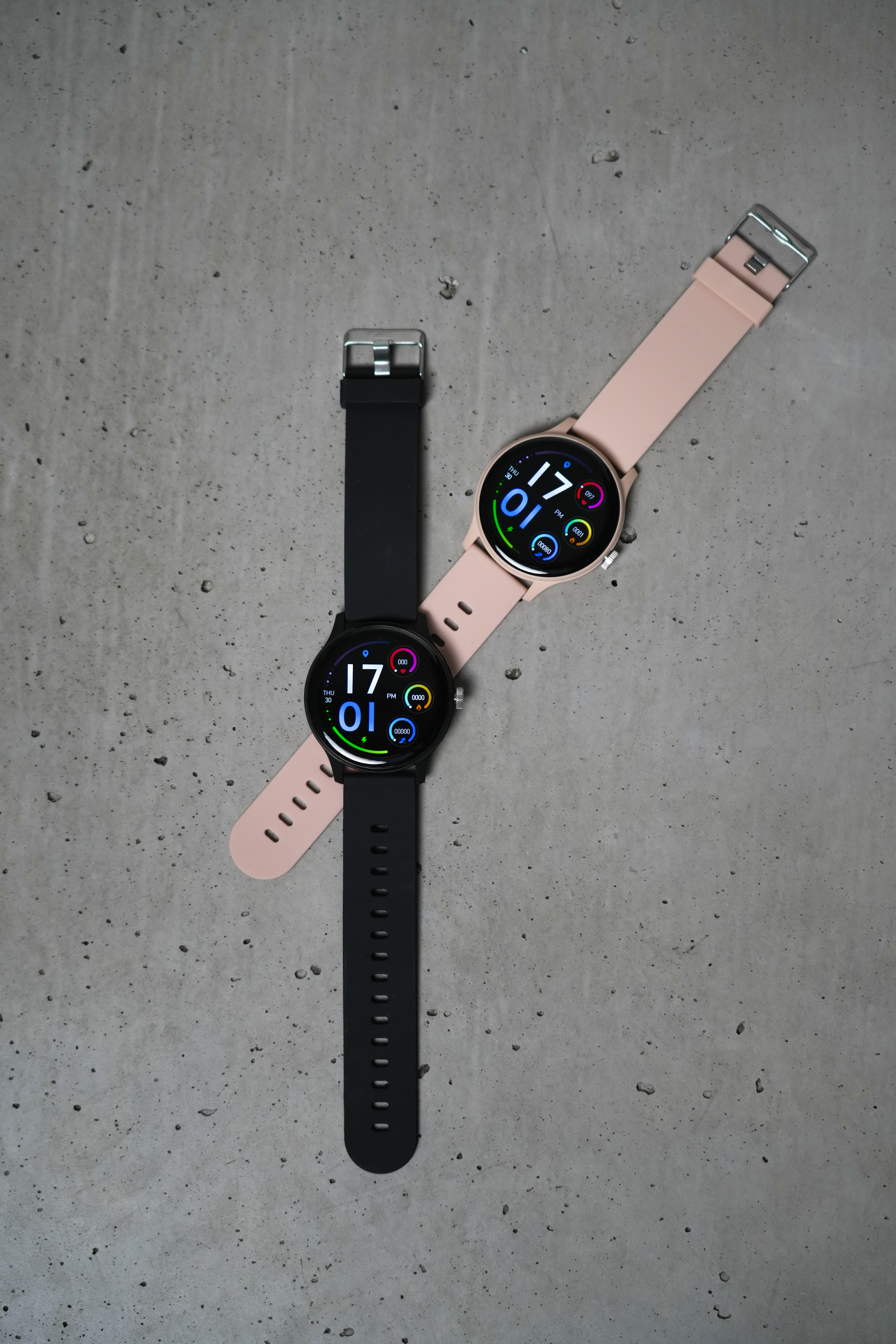 Zwei Smartwatches auf grauem Beton. Eine ist schwarz, die andere rosa. Beide zeigen die Uhrzeit 17:01.