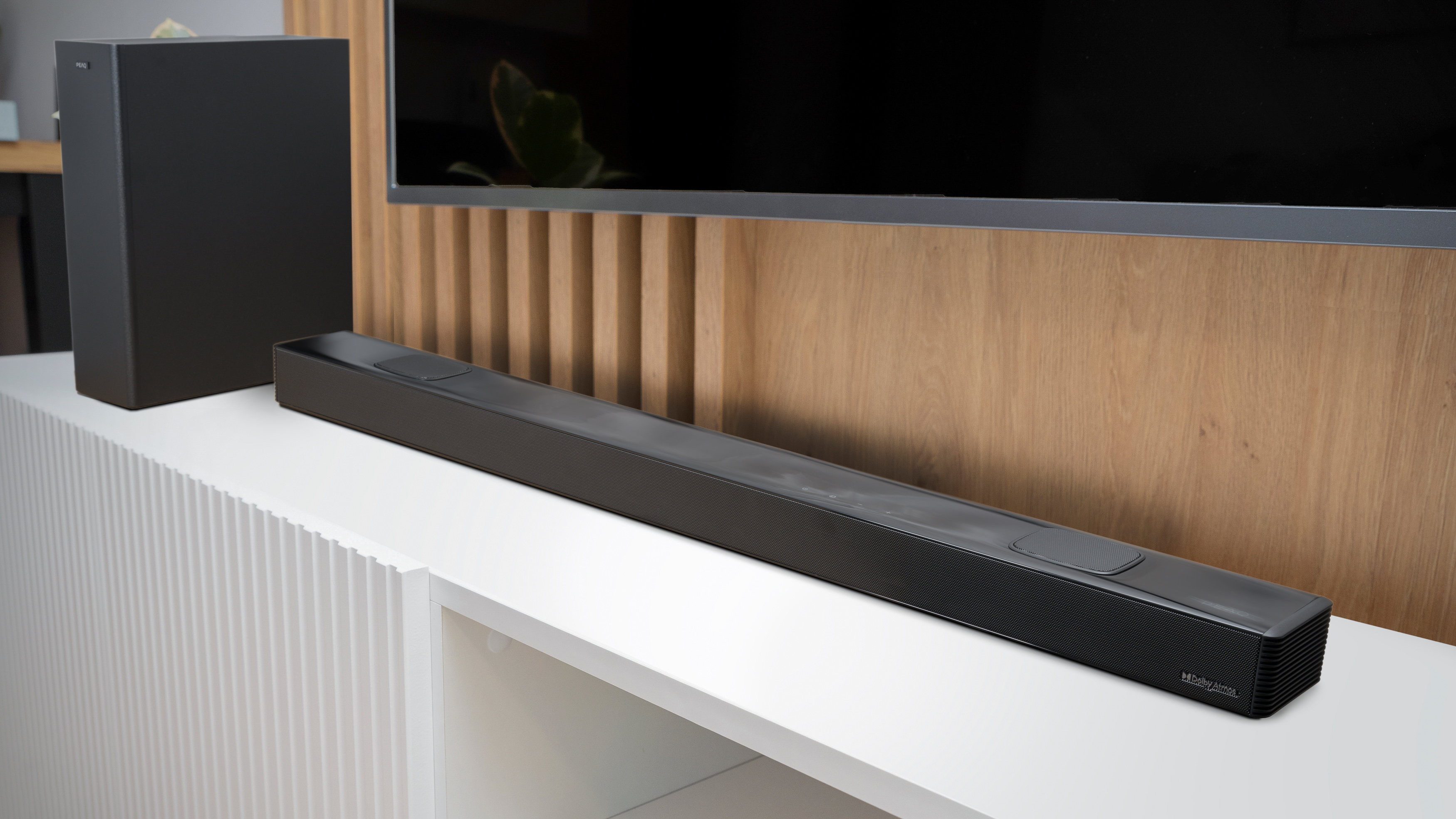 Eine schwarze Soundbar und ein Subwoofer stehen auf einer weißen Oberfläche. Dahinter befindet sich ein Fernseher über Holzvertäfelung.