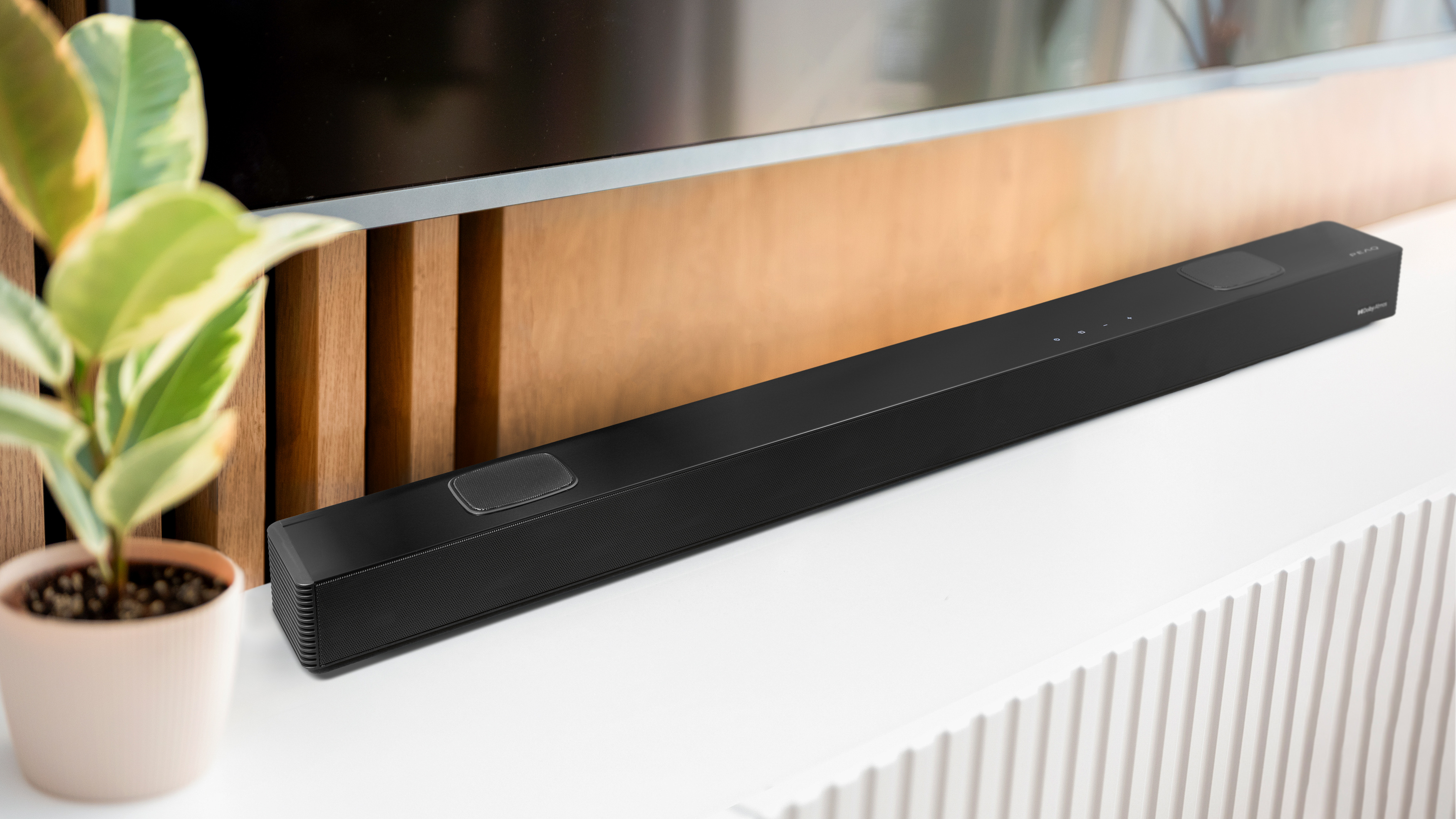 Eine schwarze Soundbar liegt auf einer weißen Oberfläche. Links ist eine Pflanze im Topf. Holzverkleidung im Hintergrund.