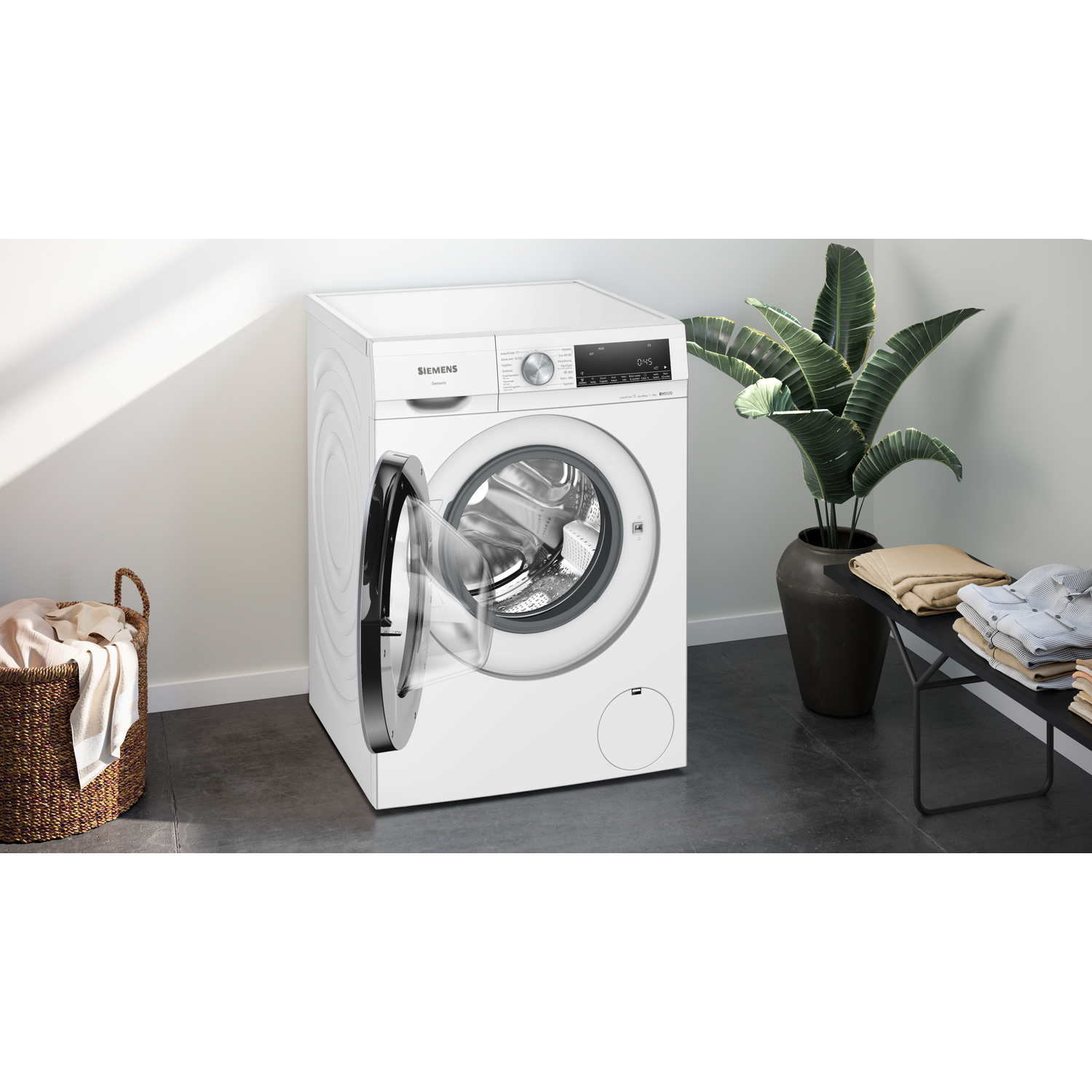 A white washing machine with an open door. A basket of clothes on the left and a plant on the right. Clothes are folded on a table.