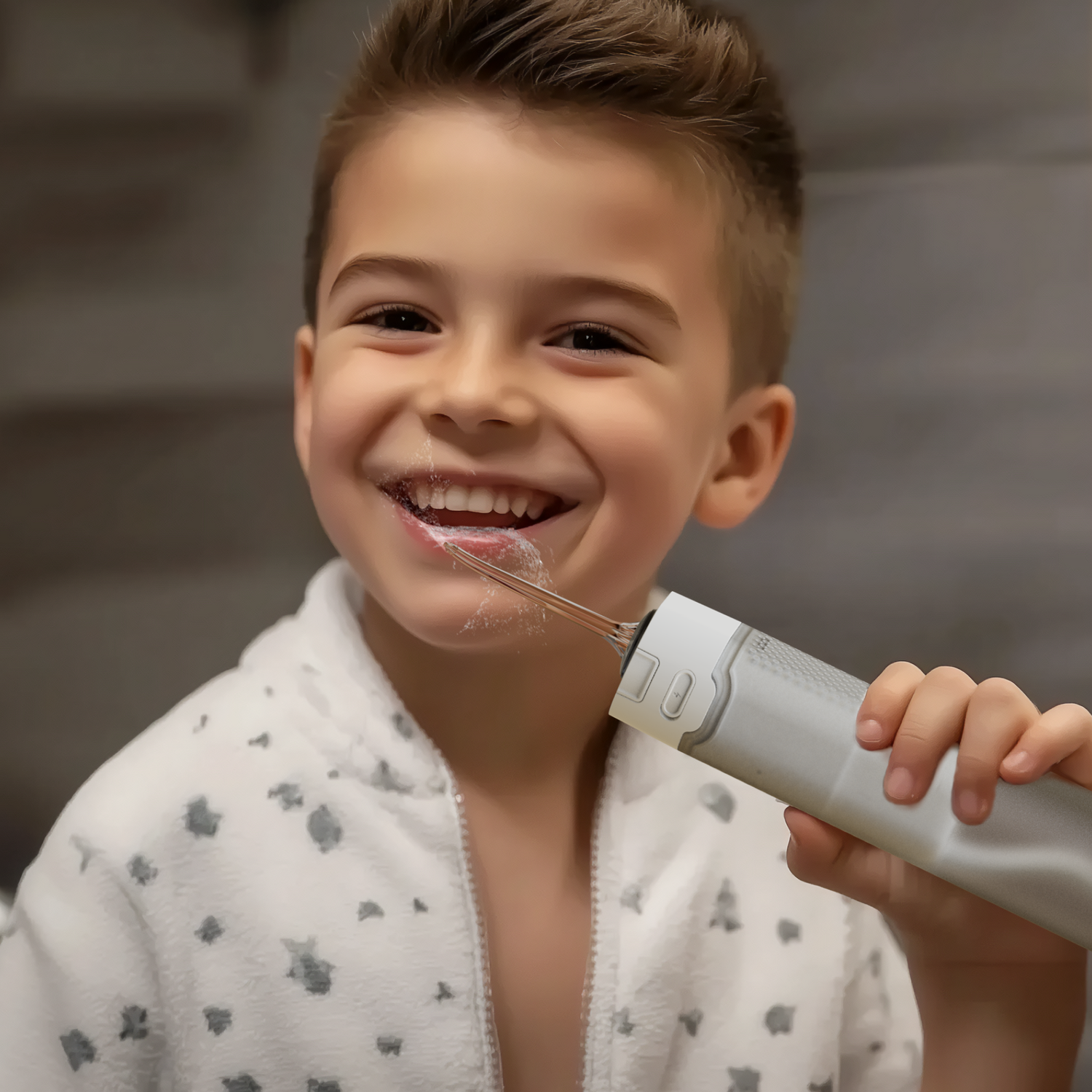 A smiling boy using a water flosser on his teeth. He wears a white robe. Background is a gray bathroom wall.
