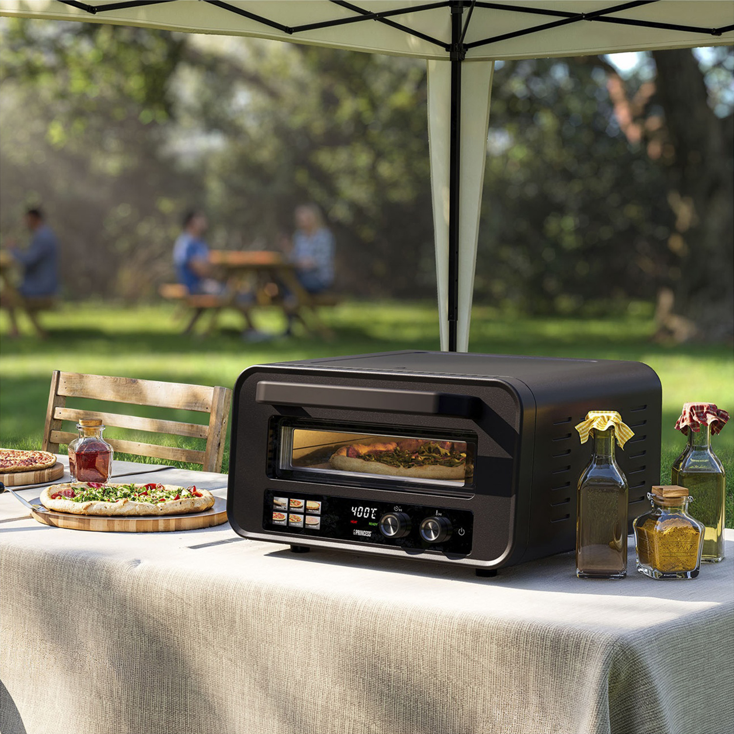 Scena all'aperto con forno per pizza, pizze, bottiglie e persone sullo sfondo. Tavolo sotto un tendone in un giardino soleggiato.