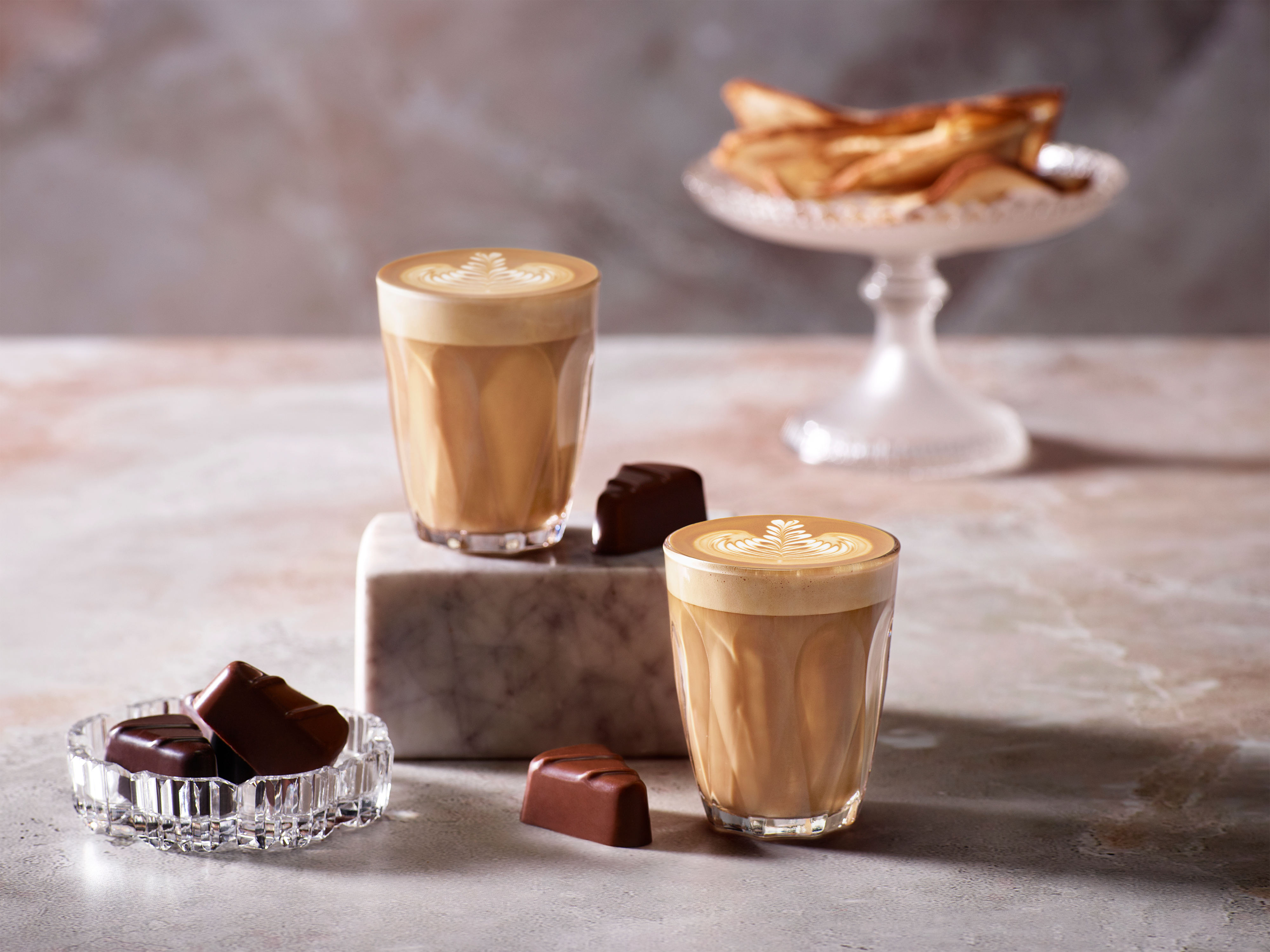Zwei Lattes mit Latte Art, Pralinen und Gebäck auf einer Marmoroberfläche. Weiche Beleuchtung.