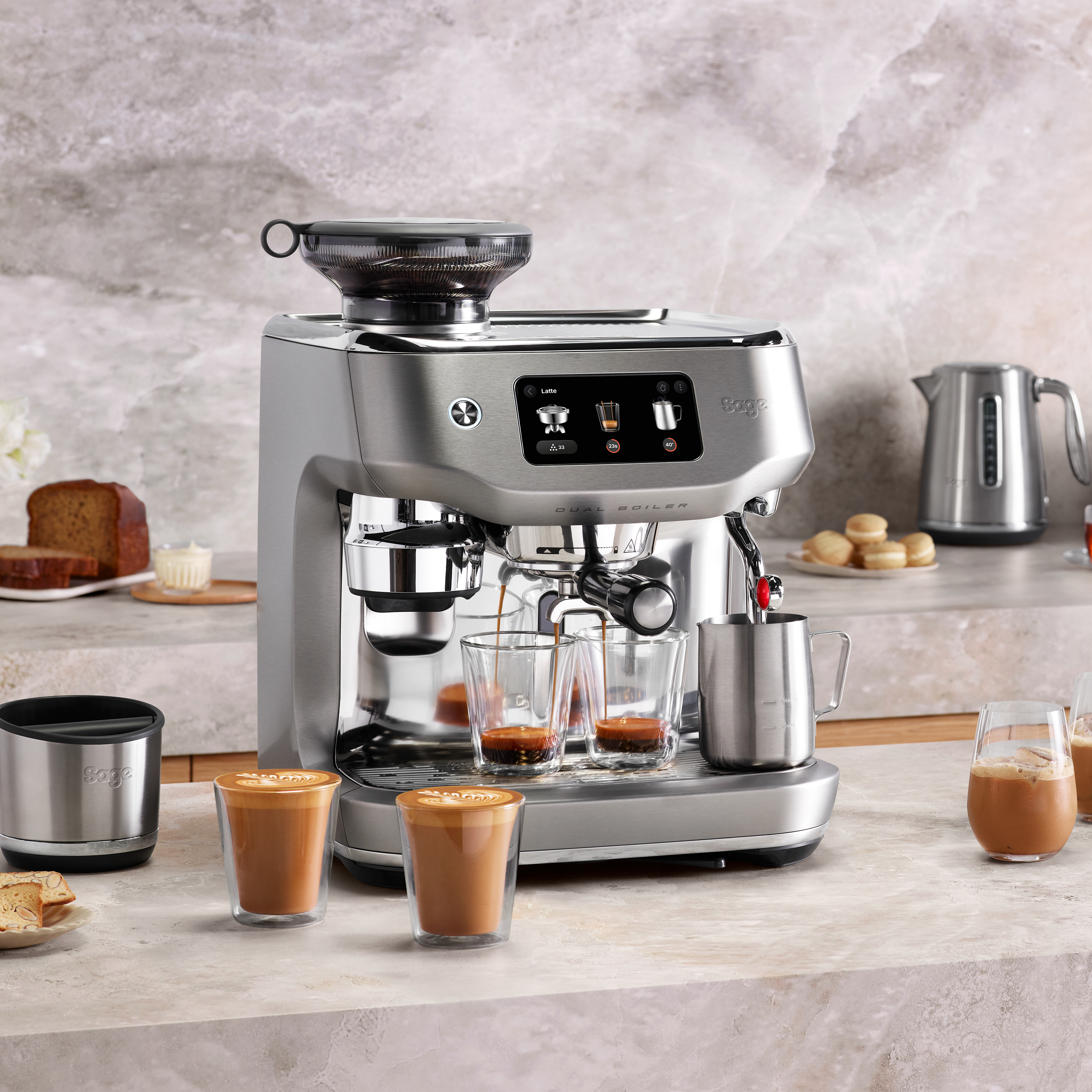 Espressomaschine mit Kaffee, Milchkännchen und Kuchen auf der Theke. Latte Art in Gläsern. Silberner Wasserkocher im Hintergrund.