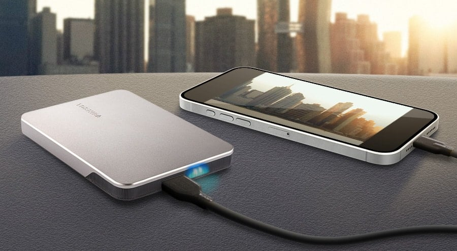 Srebrny zewnętrzny dysk twardy i smartfon połączone czarnym kablem. Rozmyty krajobraz miejski w tle.