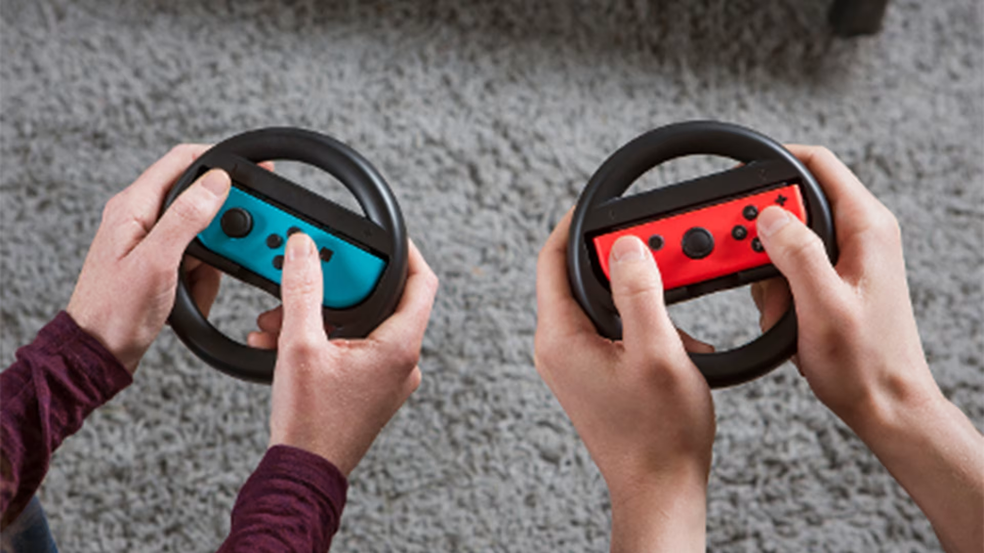 Twee handen die gamecontrollers vasthouden in stuurwielbevestigingen. Eén is blauw, de andere rood, op een grijs tapijt.
