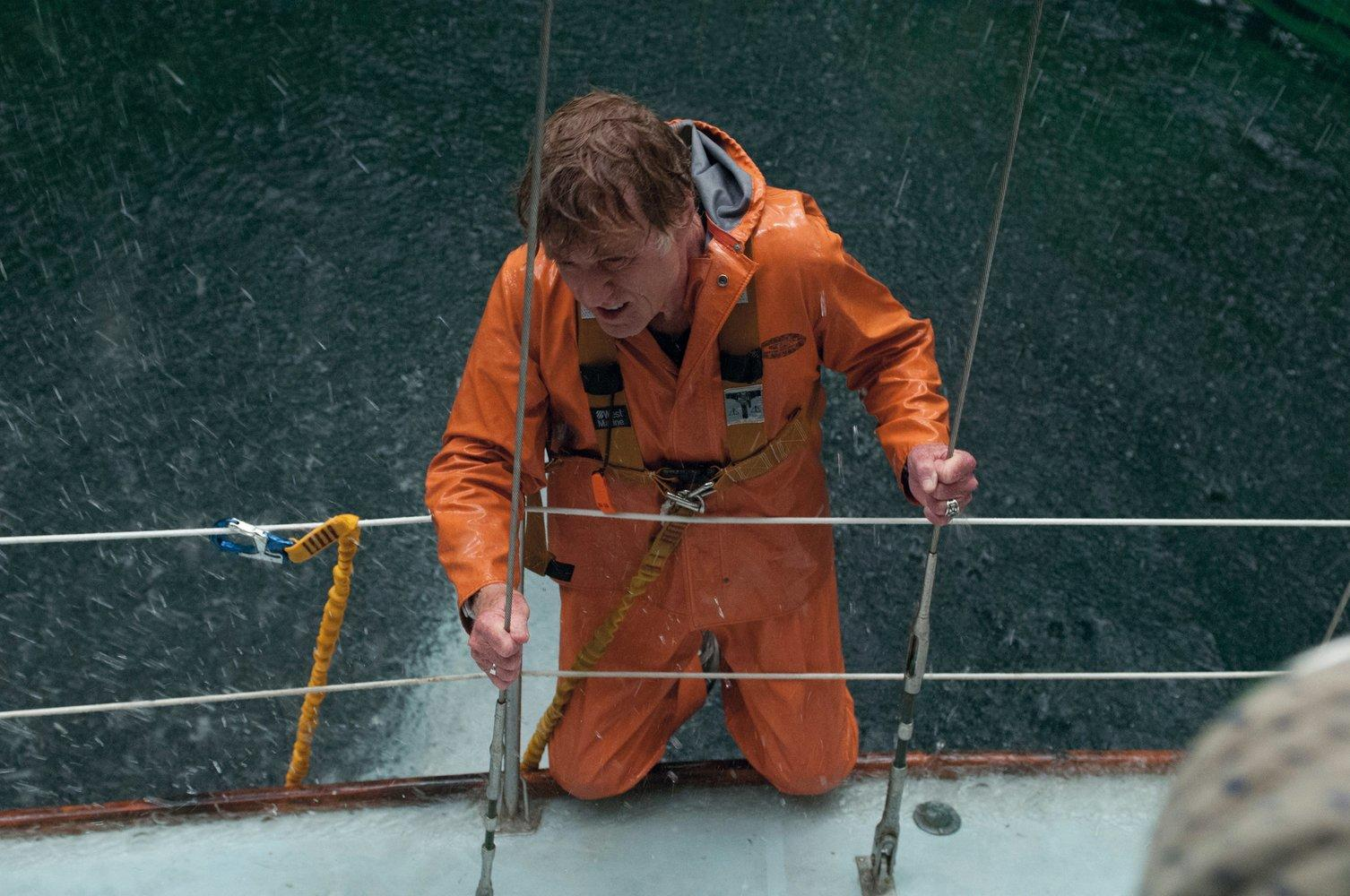 Ein Mann in einem orangefarbenen Anzug auf einem Boot in einem Sturm. Er hält sich am Geländer fest. Starker Regen fällt um ihn herum.