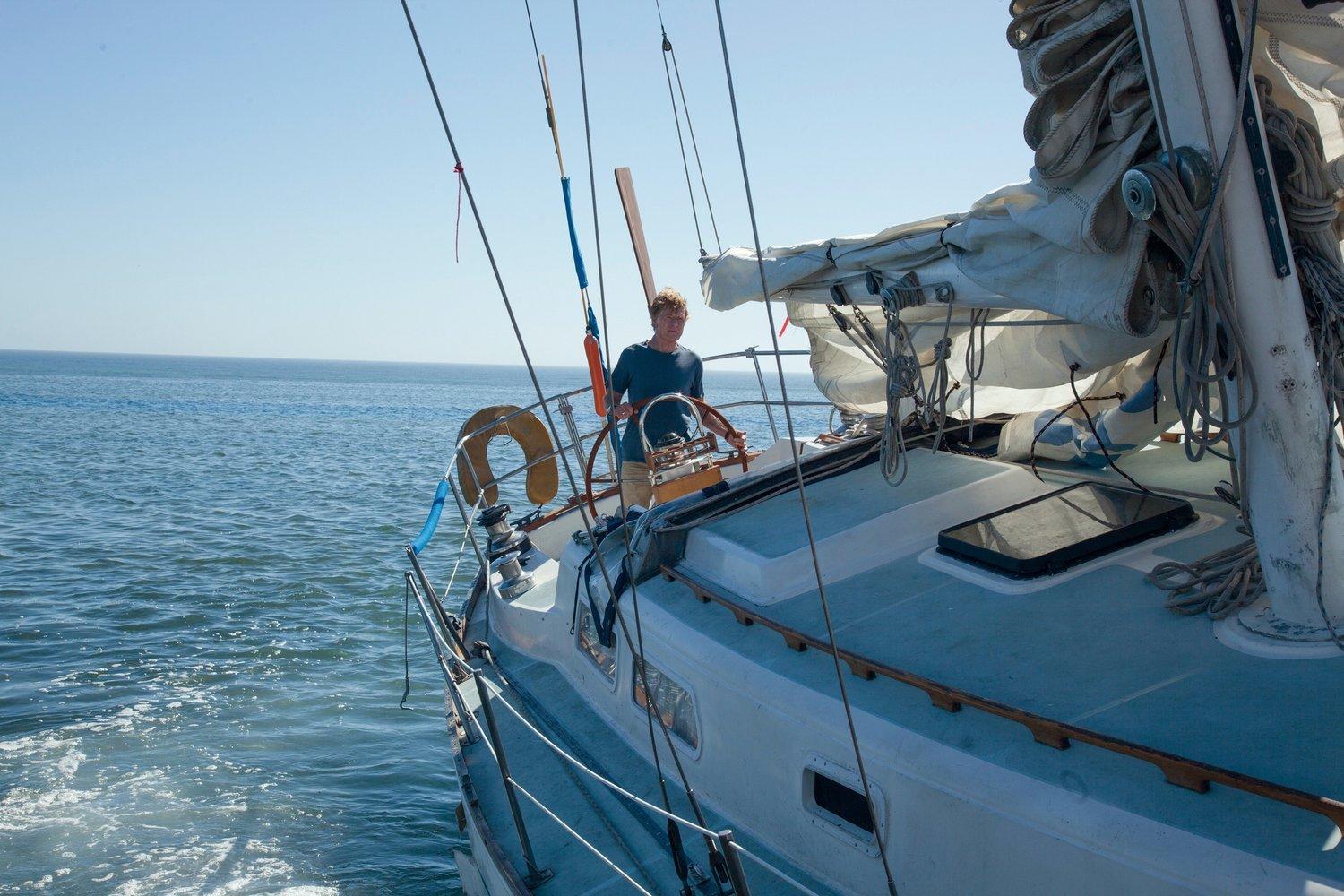 Ein Mann steuert an einem sonnigen Tag ein Segelboot. Das Boot ist weiß, blaues Wasser umgibt es. Der Himmel ist klar.