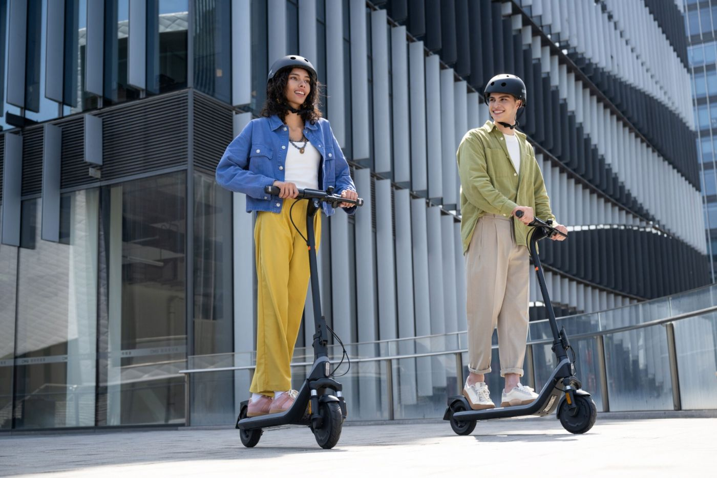 Két ember robogón, sisakot viselve. Az egyik kék dzsekiben, sárga nadrágban, a másik zöld ingben, bézs nadrágban, egy épület közelében.