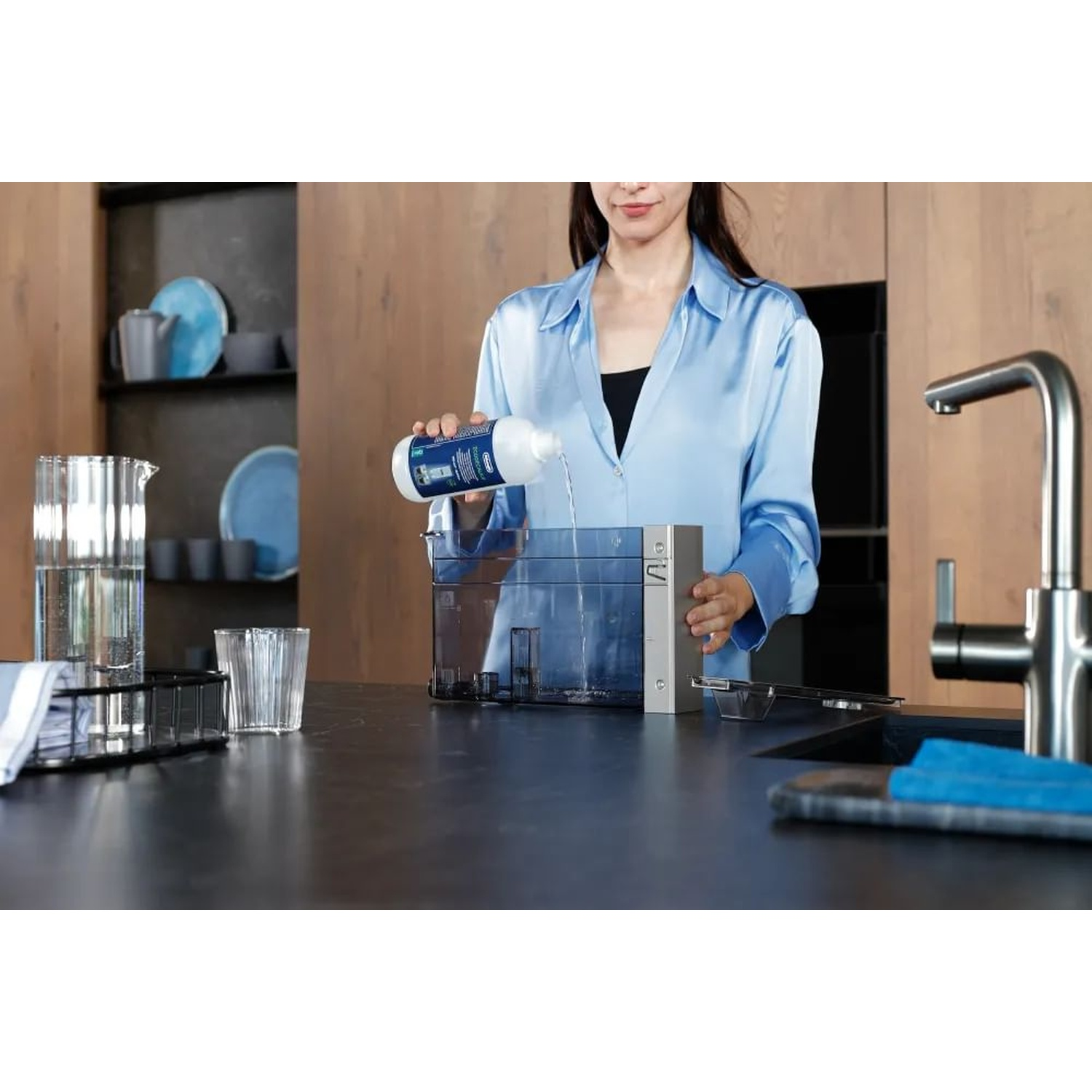 Una donna con una camicia blu versa del liquido da una bottiglia in un serbatoio d'acqua. Ambiente cucina con elettrodomestici e accessori.