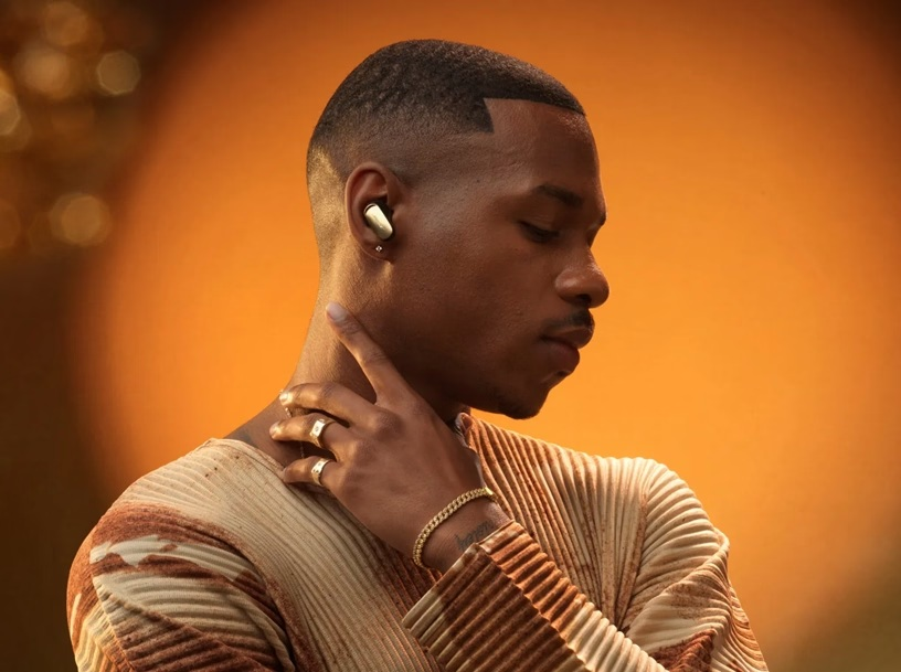 Mężczyzna z krótkimi włosami w słuchawkach, biżuterii i koszuli z fakturą dotyka palcem szyi.