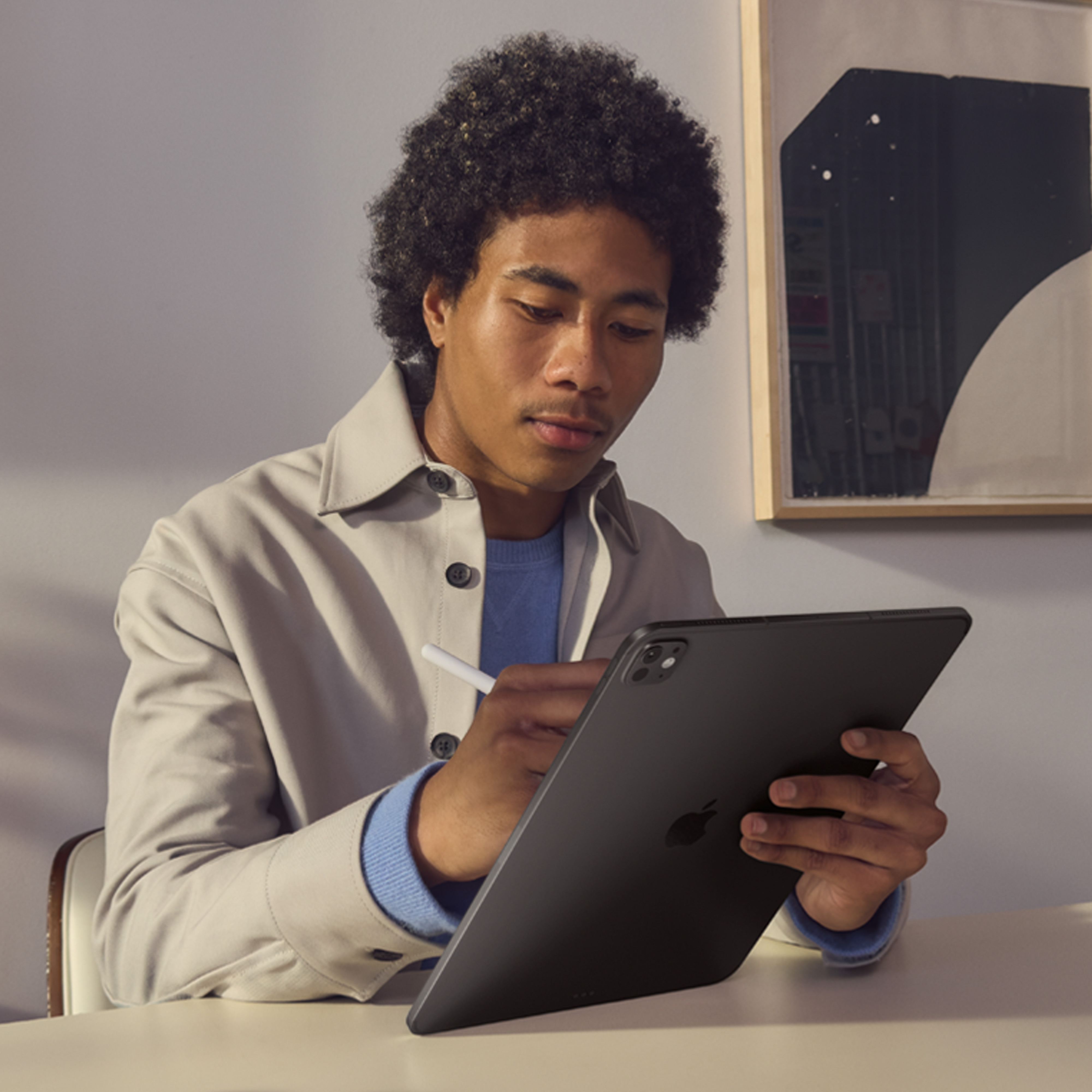 Un uomo con i capelli afro usa un tablet e una penna su una scrivania. Indossa un cappotto chiaro e un maglione blu.