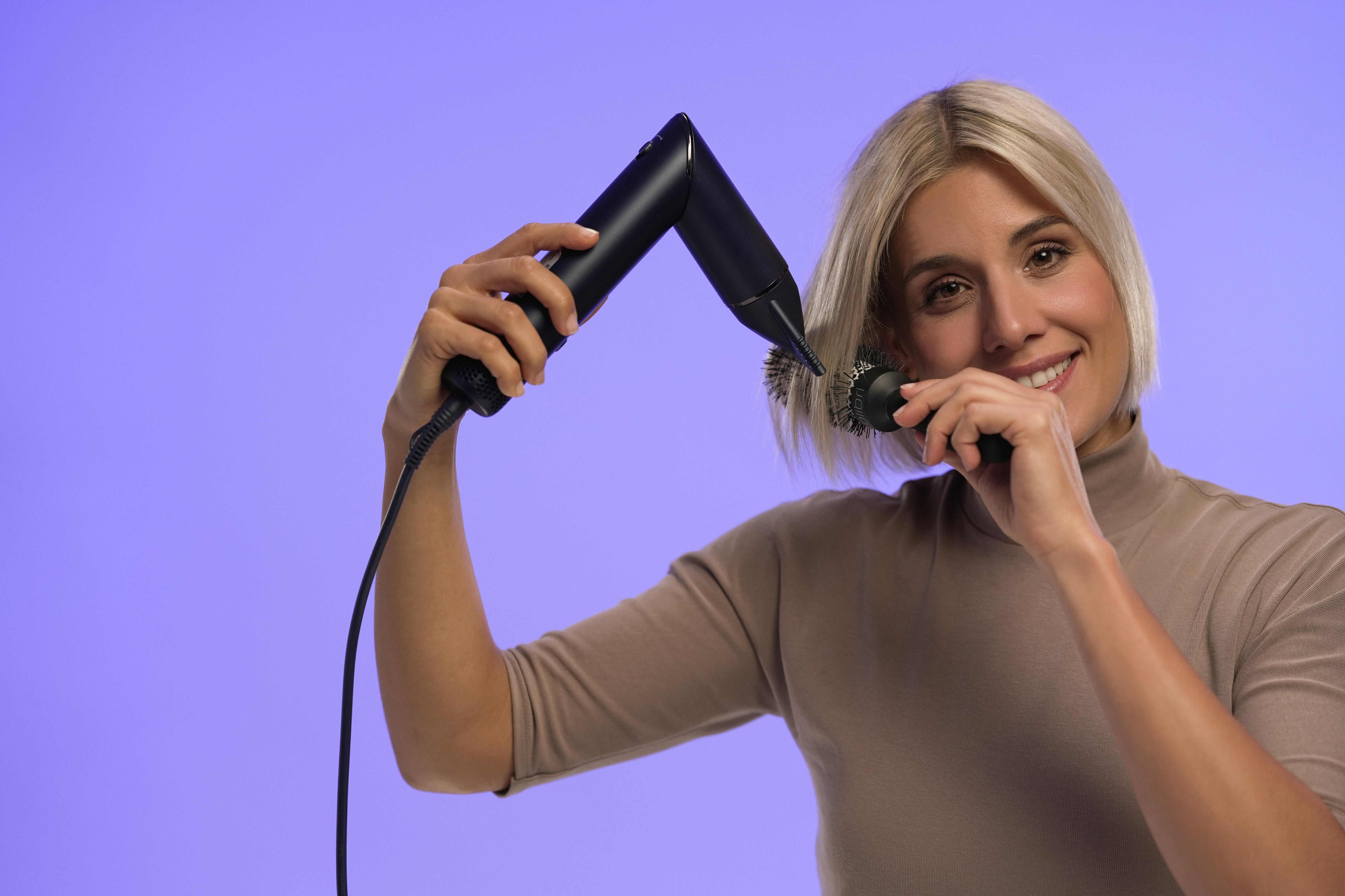 A woman with blonde hair smiles, holding a hair dryer and a round brush, against a purple background.