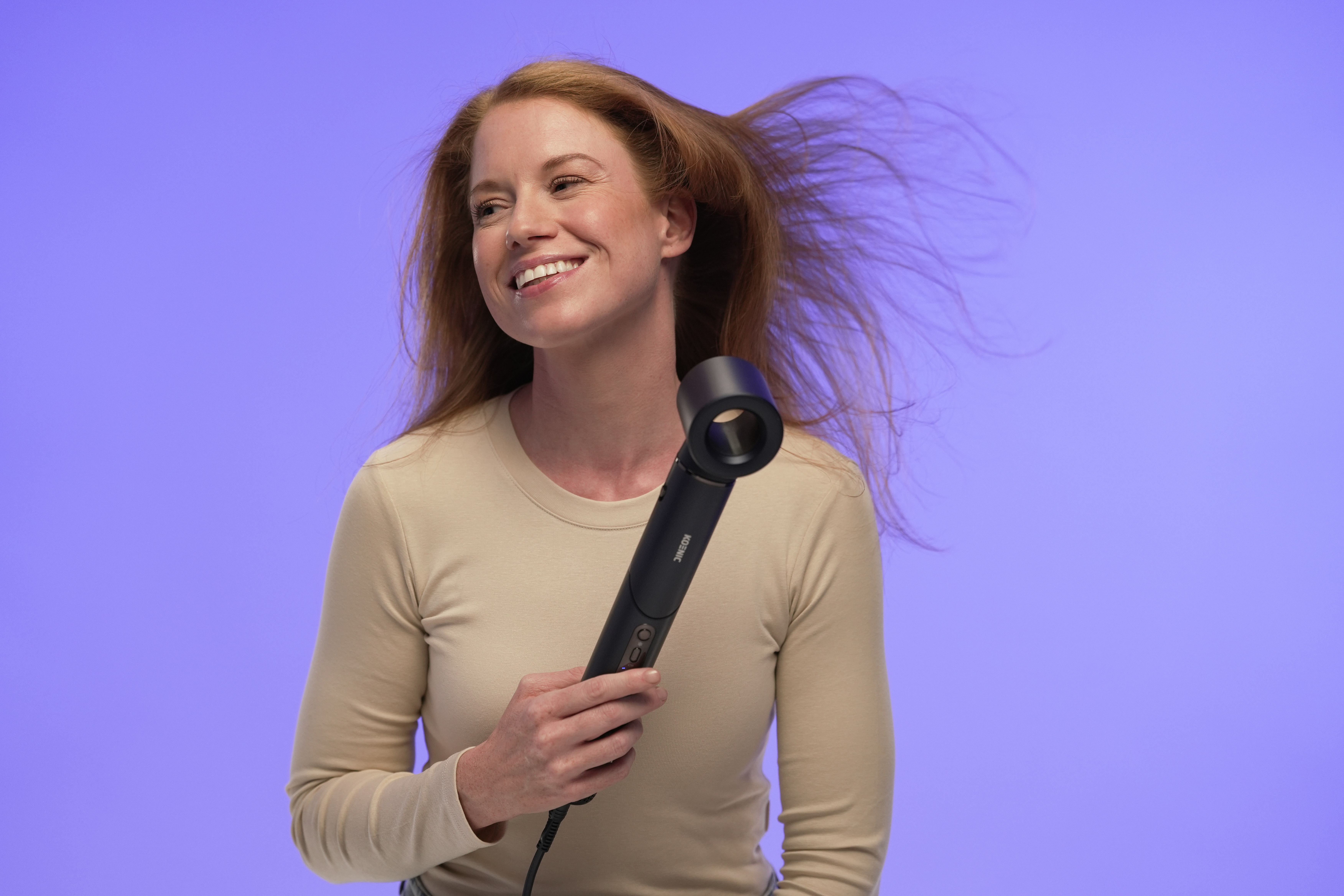 A smiling woman with red hair holds a black hair dryer against a purple background, her hair blowing.
