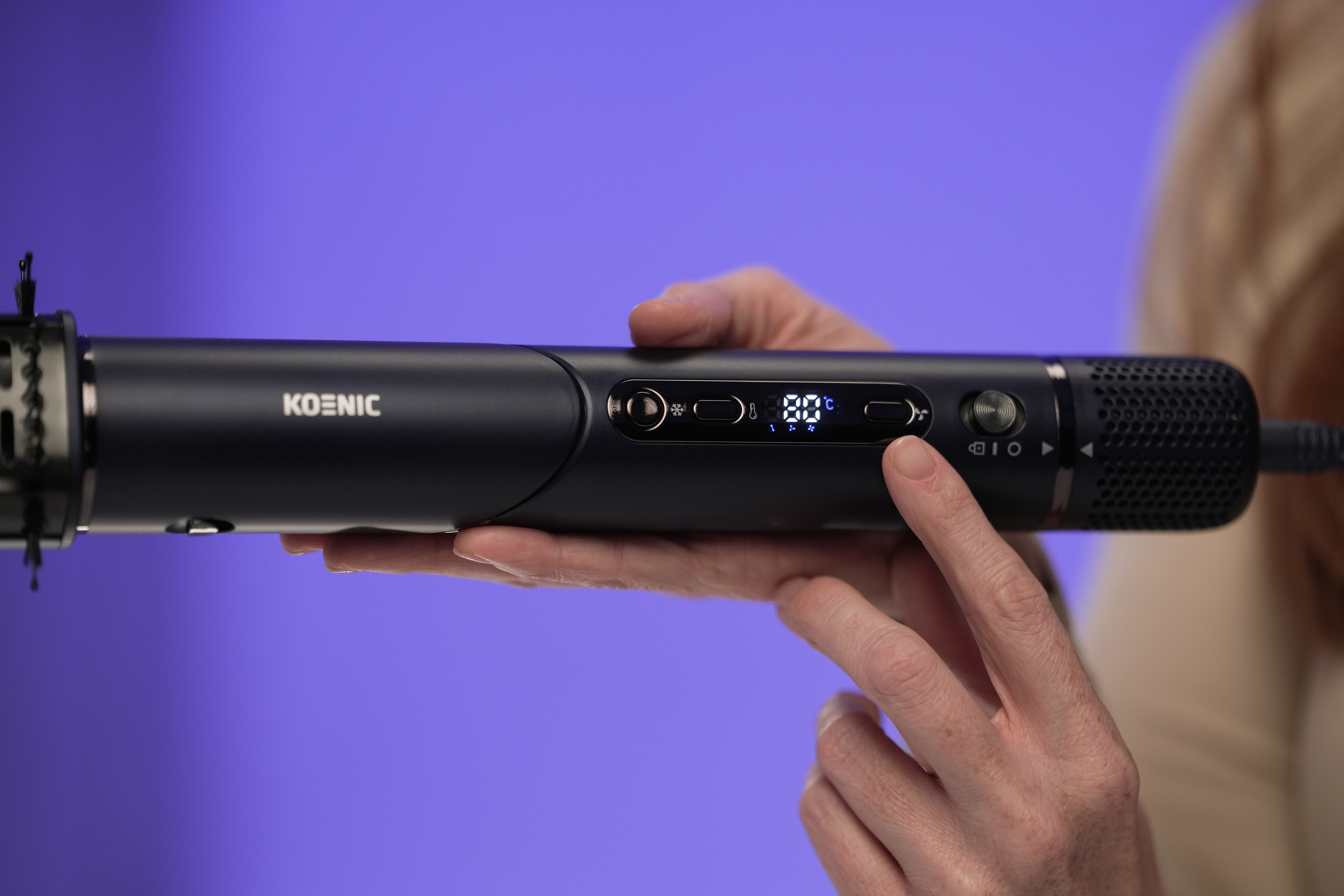 A person holds a black hair styling tool. The tool displays a temperature of 80 degrees. Purple background.