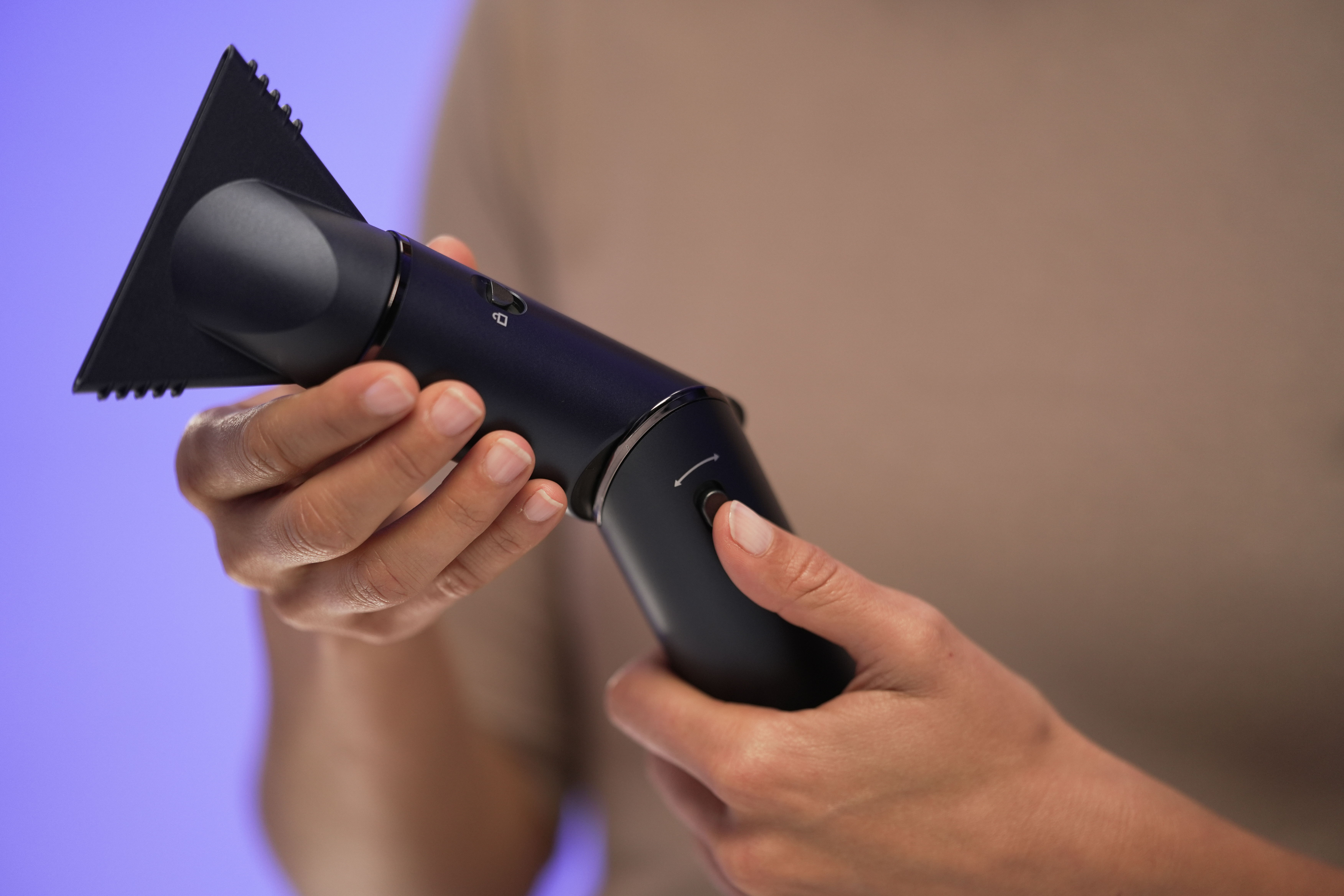 Hands holding a dark blue hair tool. It has a triangular nozzle and several buttons. Background is a gradient blue to brown.