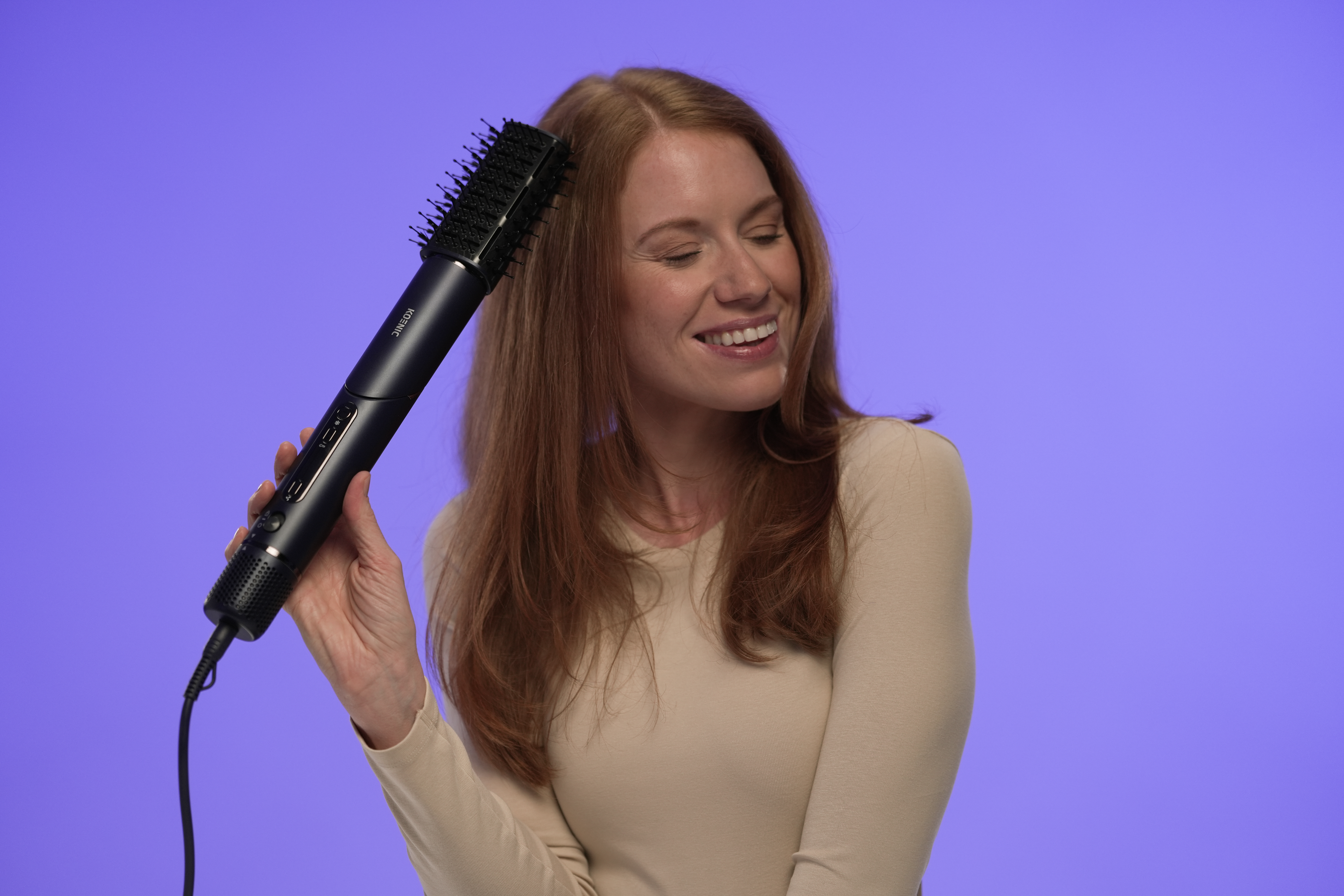 A woman with red hair smiles while holding a black hair styling tool. The background is purple.