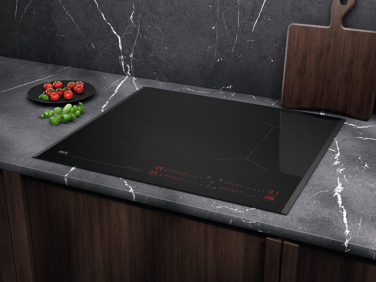 Una placa de inducción negra con pantalla roja está sobre una encimera. Hay tomates y albahaca en un plato. Hay una tabla de cortar de madera.