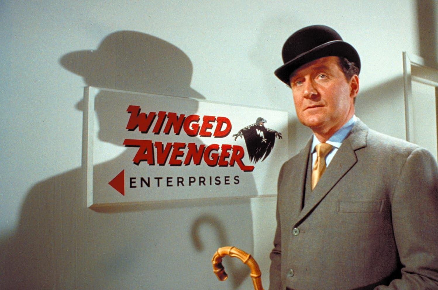 Ein Mann im Anzug und mit Melone steht neben einem Schild mit der Aufschrift 'Winged Avenger Enterprises'.