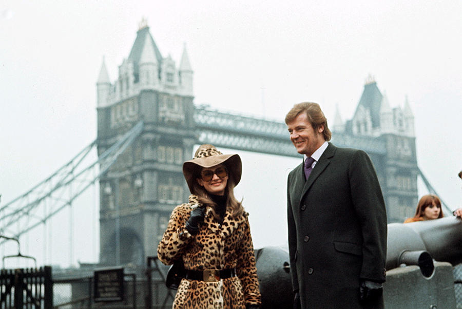 Ein Mann und eine Frau vor der Tower Bridge. Die Frau trägt einen Leopardenmantel und -hut. Der Mann trägt einen Anzug.