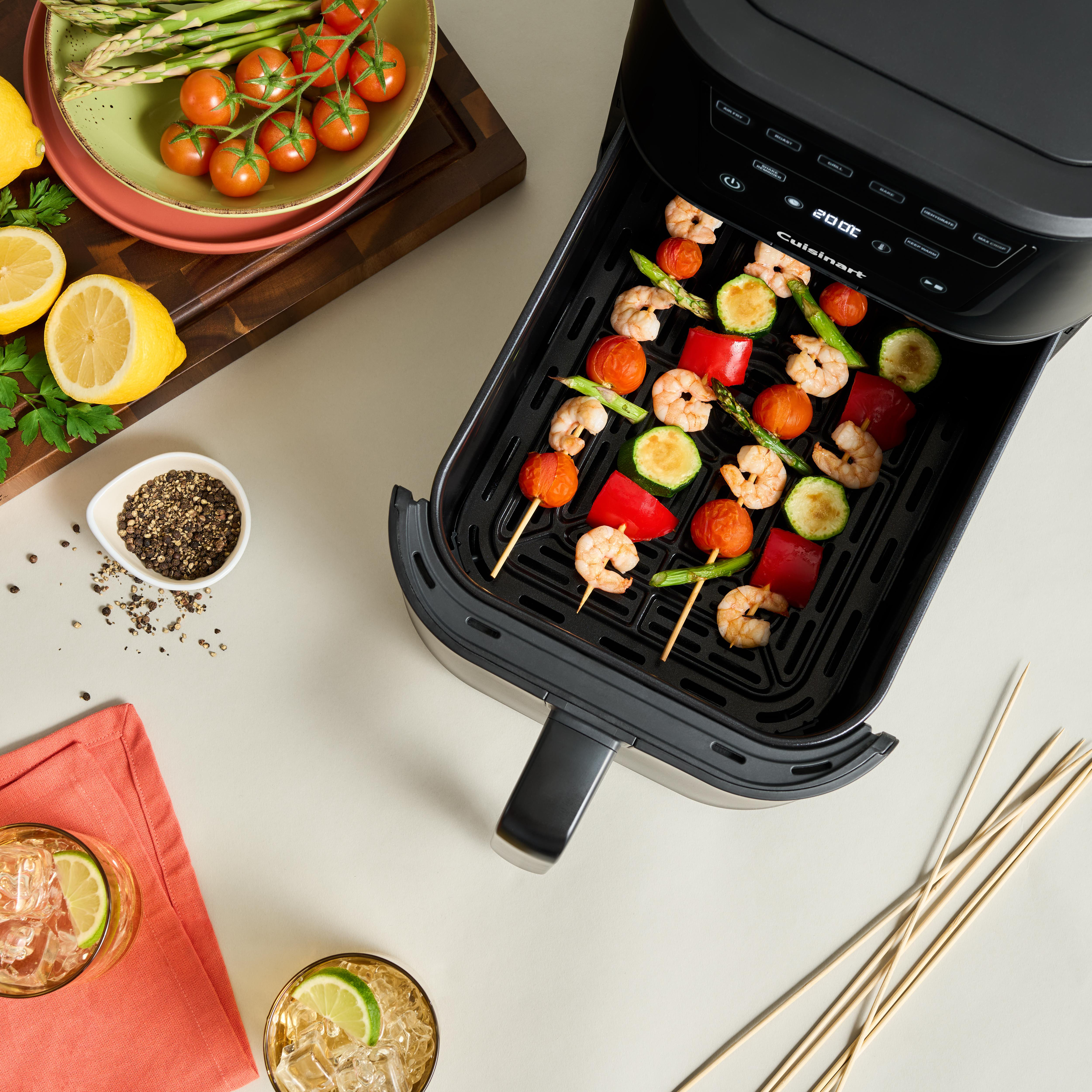 Ein Airfryer mit Spießen aus Garnelen, Tomaten und Gemüse. Zitronen, Tomaten und Getränke sind ebenfalls vorhanden.