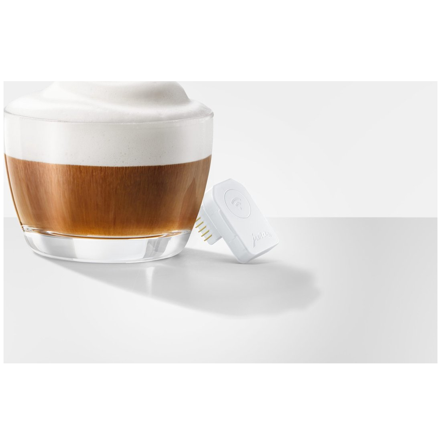 A glass of coffee with milk foam, a white wireless device with pins, on a white surface.