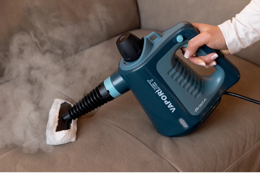 Egy személy VAPORJET-tel gőzöl egy barna bőr kanapét. A gőz a tisztítófejből áramlik.
