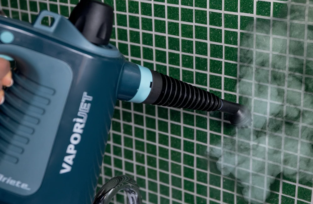Egy türkizkék VAPORJET gőzölő zöld csempézett falat tisztít. A fúvókából gőz áramlik ki. Egy kéz tartja a gőzölőt.