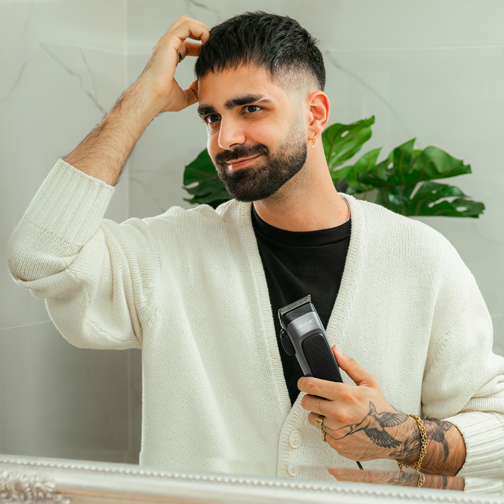 Un hombre con barba, sosteniendo una cortadora de pelo, mirándose en el espejo. Viste una rebeca blanca y tiene tatuajes.
