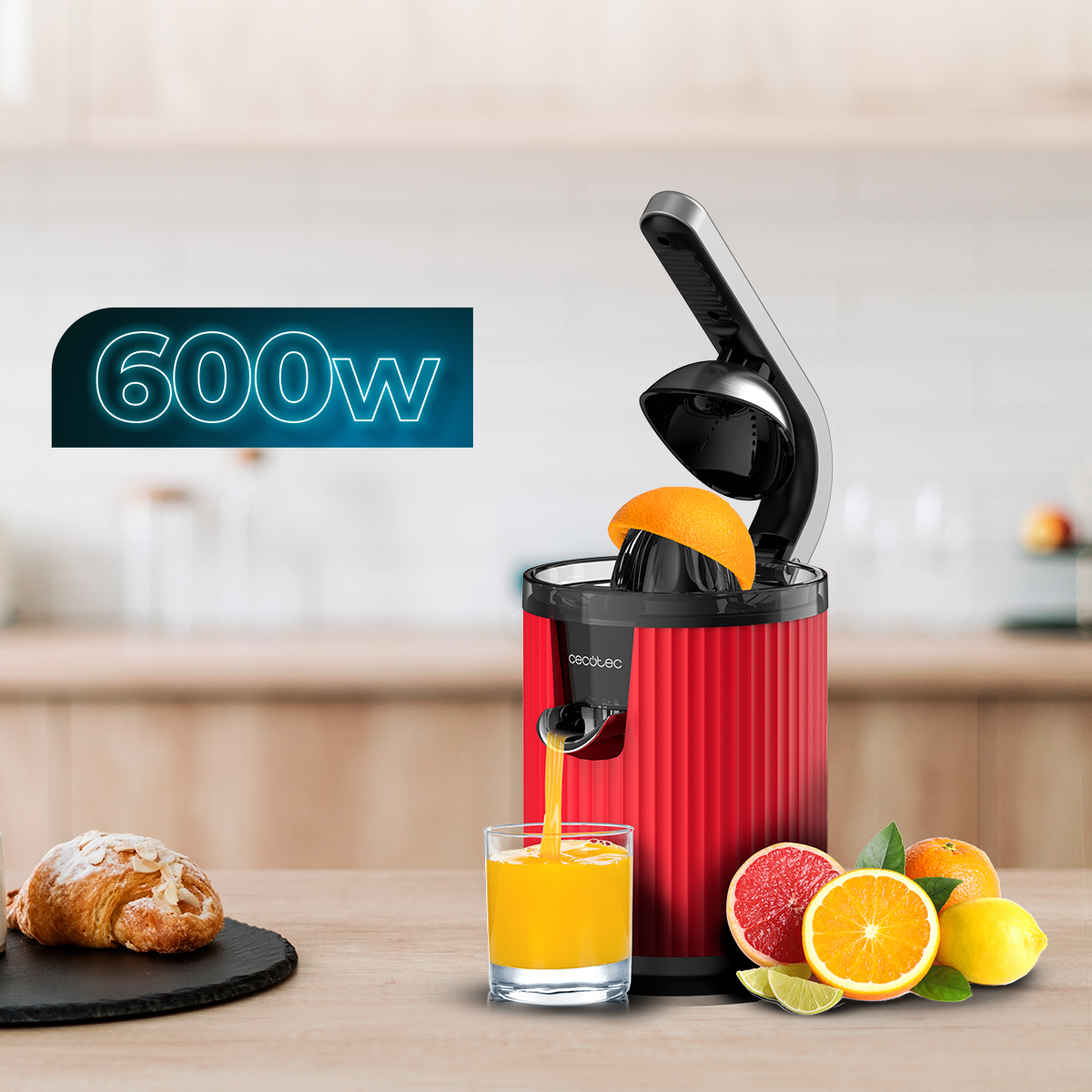 Exprimidor rojo con naranja. Vaso de zumo, cítricos y croissant sobre una mesa. Cartel de 600W al fondo.