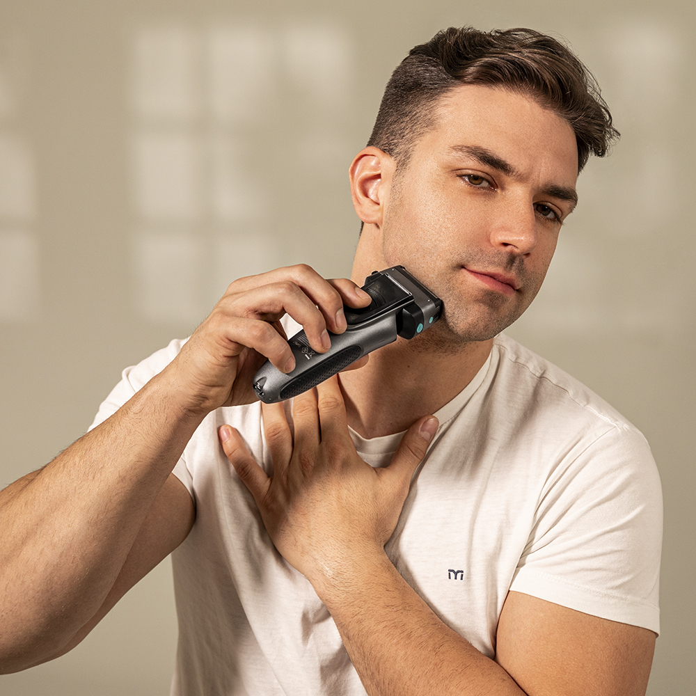 Un hombre se afeita la barba con una afeitadora eléctrica. Lleva una camiseta blanca. El fondo es de color beige liso.