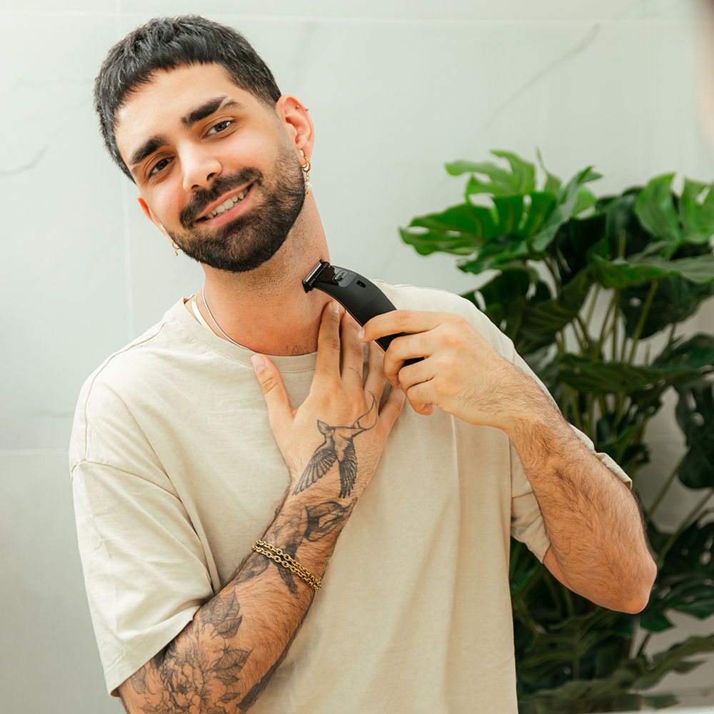 Un hombre se afeita el cuello con una recortadora eléctrica. Tiene tatuajes, una cadena de oro y pendientes. Una planta y un espejo están en el fondo.