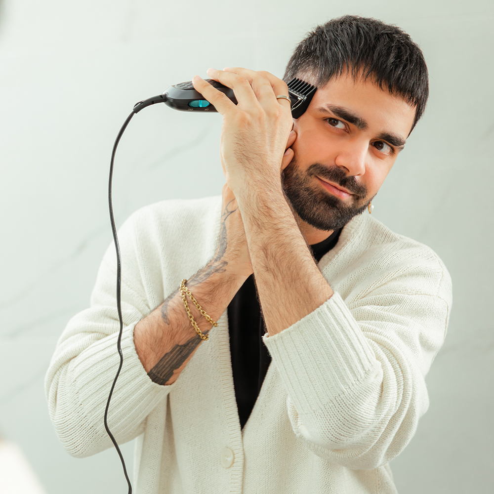 Un hombre con barba usa una cortadora de pelo en su cabeza. Lleva un suéter blanco y tiene un tatuaje en el brazo.