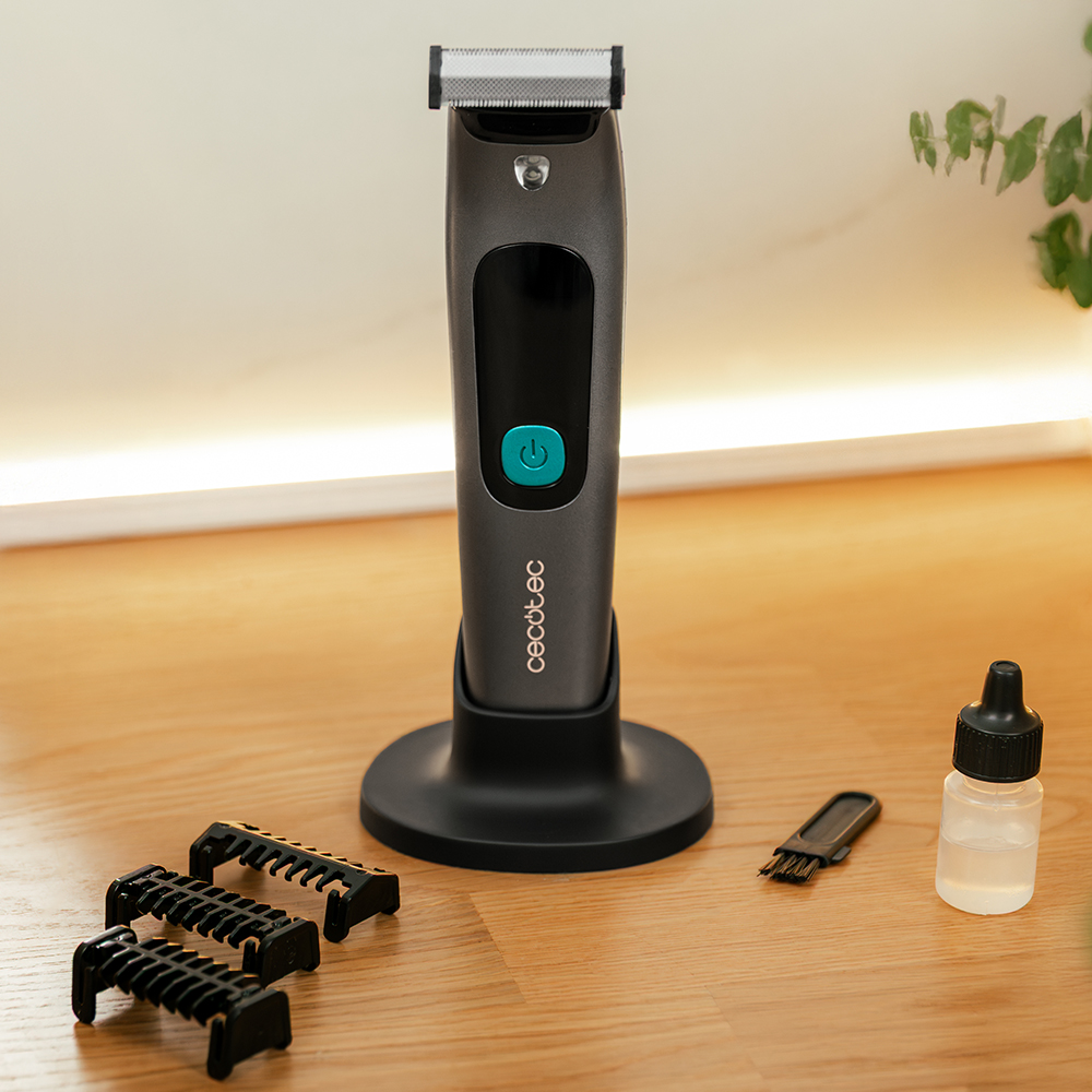 Recortadora eléctrica con accesorios sobre una superficie de madera. Incluye cuchillas, cepillo de limpieza y frasco de aceite. Recortadora gris con botón verde.