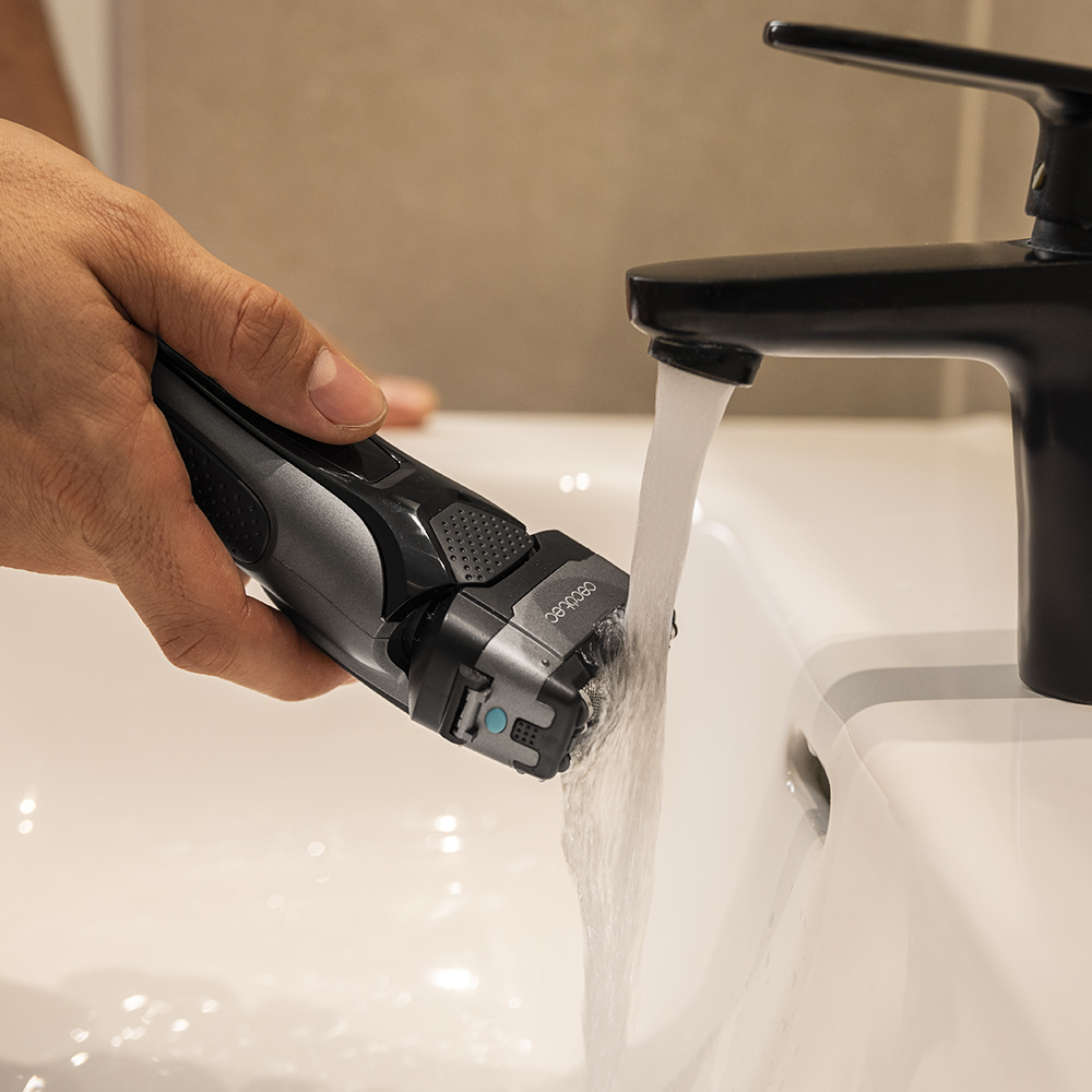 Una mano sostiene una afeitadora eléctrica gris bajo agua corriente de un grifo negro en un lavabo blanco.