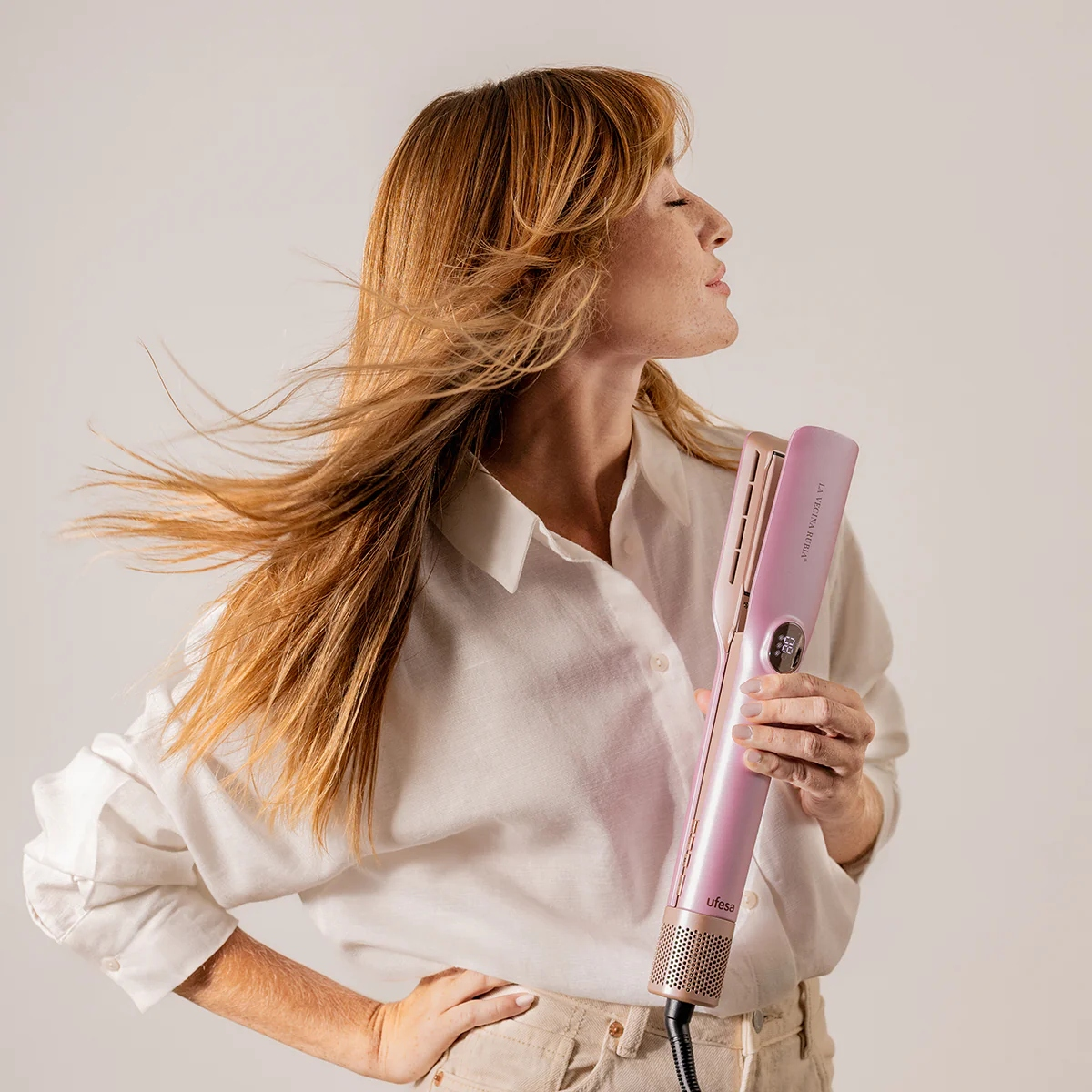 Mujer con cabello rojo ondeante sostiene una plancha de pelo rosa. Viste camisa blanca y pantalones beige, posando sobre fondo blanco.