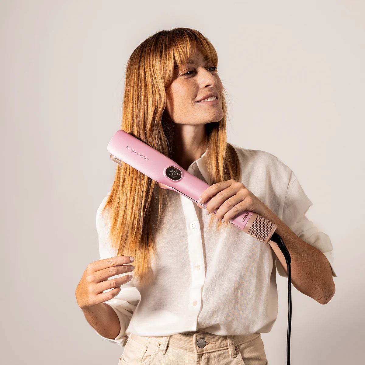 Mujer pelirroja usando una plancha rosa. Lleva camisa blanca y pantalones beiges. Fondo blanco.
