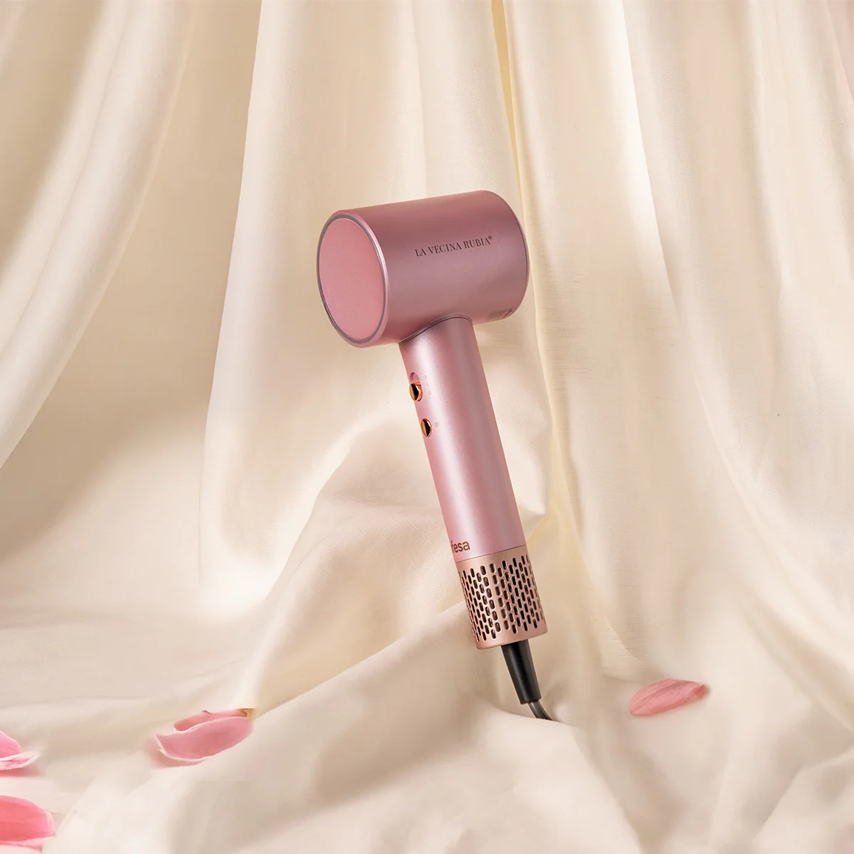 Un secador de pelo rosa sobre una tela crema. Pétalos esparcidos. El secador tiene la frase 'LA VECINA RUBIA'.
