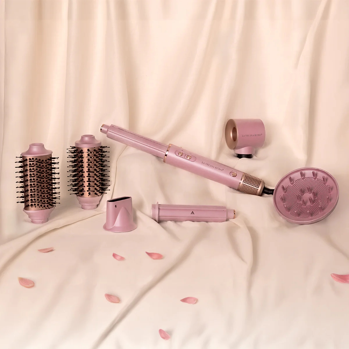 Un conjunto rosa para peinar el cabello está dispuesto sobre una tela color crema. Incluye un secador de pelo, accesorios de cepillo y un difusor, con pétalos esparcidos.