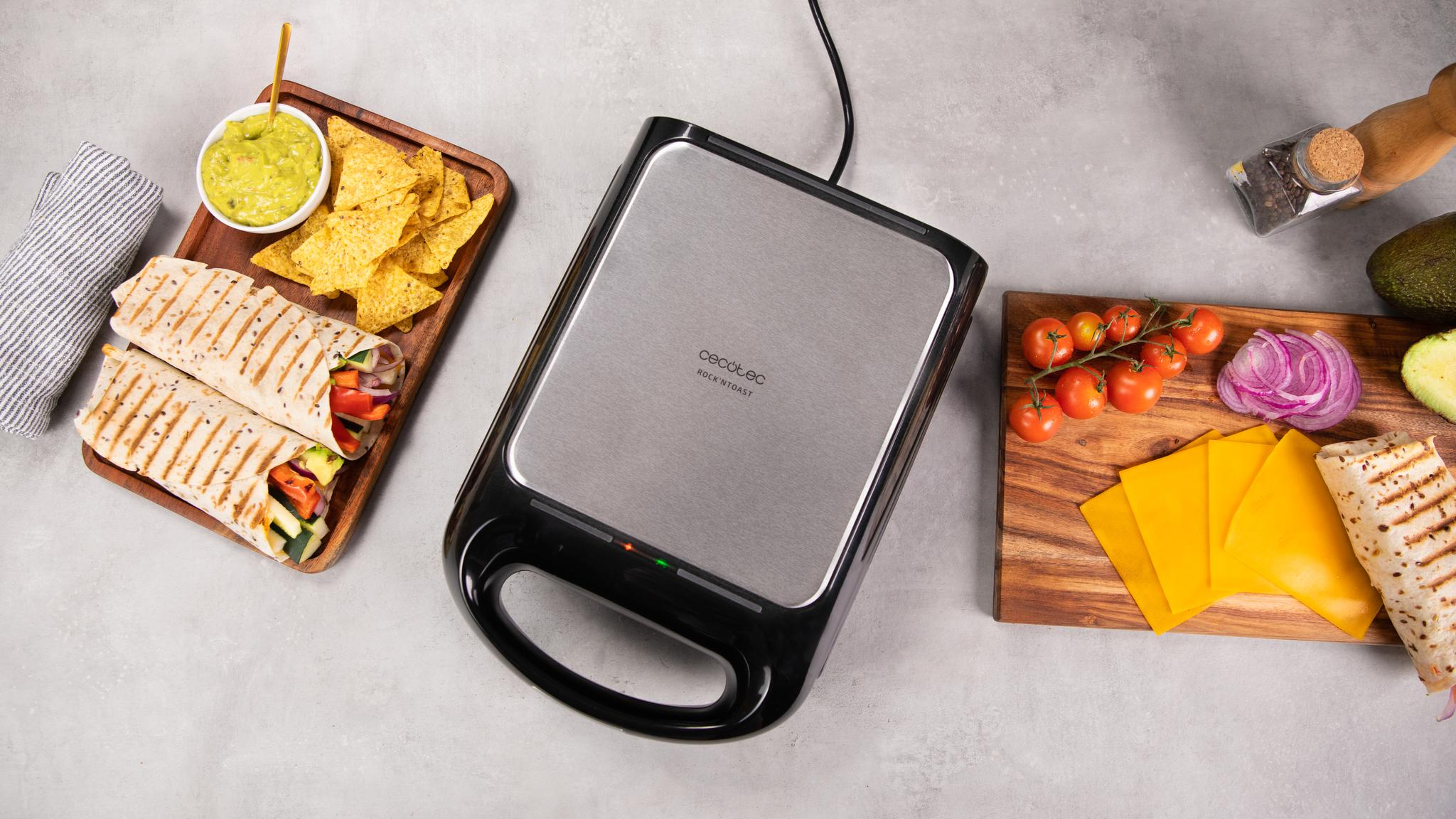 Una sandwichera con un sándwich e ingredientes. Tortillas, guacamole, patatas fritas, tomates, queso y cebolla están en la mesa.
