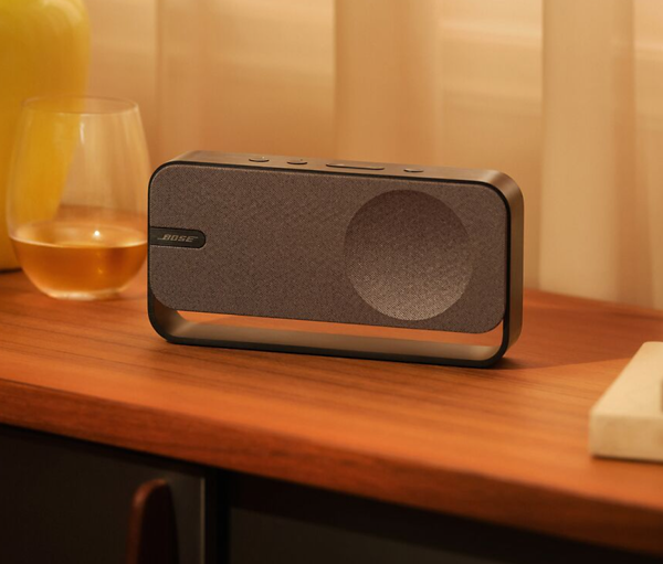 Un altavoz Bose está sobre una superficie de madera junto a un vaso con líquido. Un marco metálico curvado rodea el altavoz. Iluminación suave.